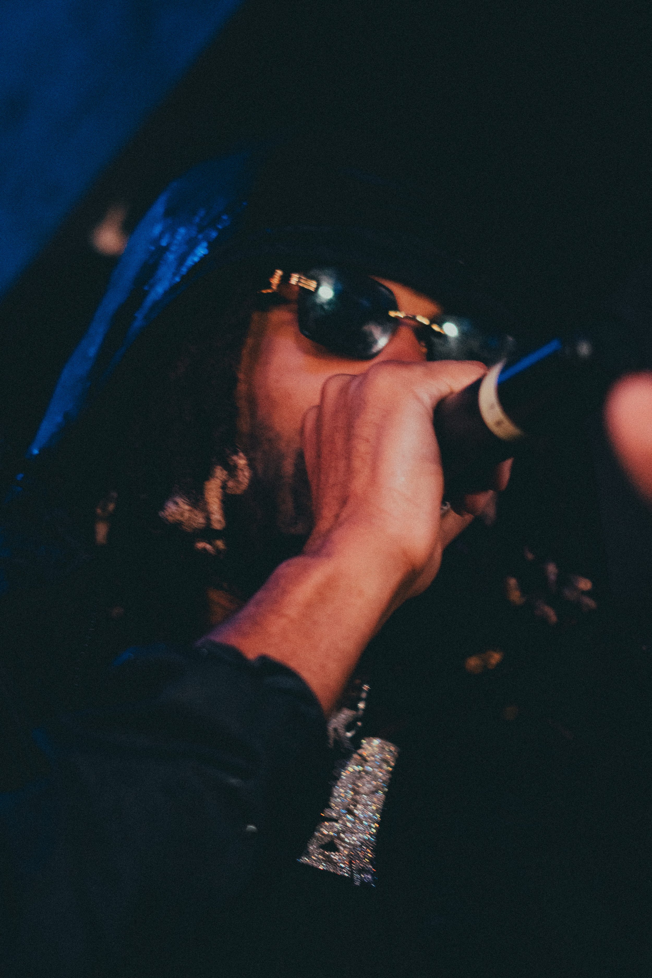 Une main tenant un cigare dans une scène sombre, avec une personne portant des lunettes de soleil et un chapeau en arrière-plan, en ambiance nocturne