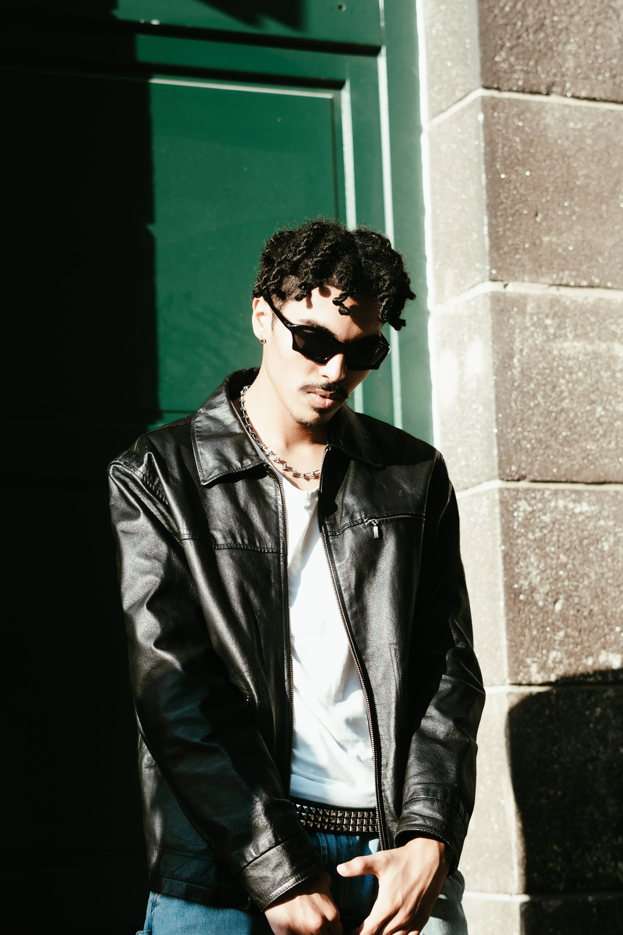 Jeune homme portant des lunettes de soleil, une veste en cuir noir et un t-shirt blanc, se tenant près d'un mur de pierre avec une porte verte derrière lui, en plein soleil.