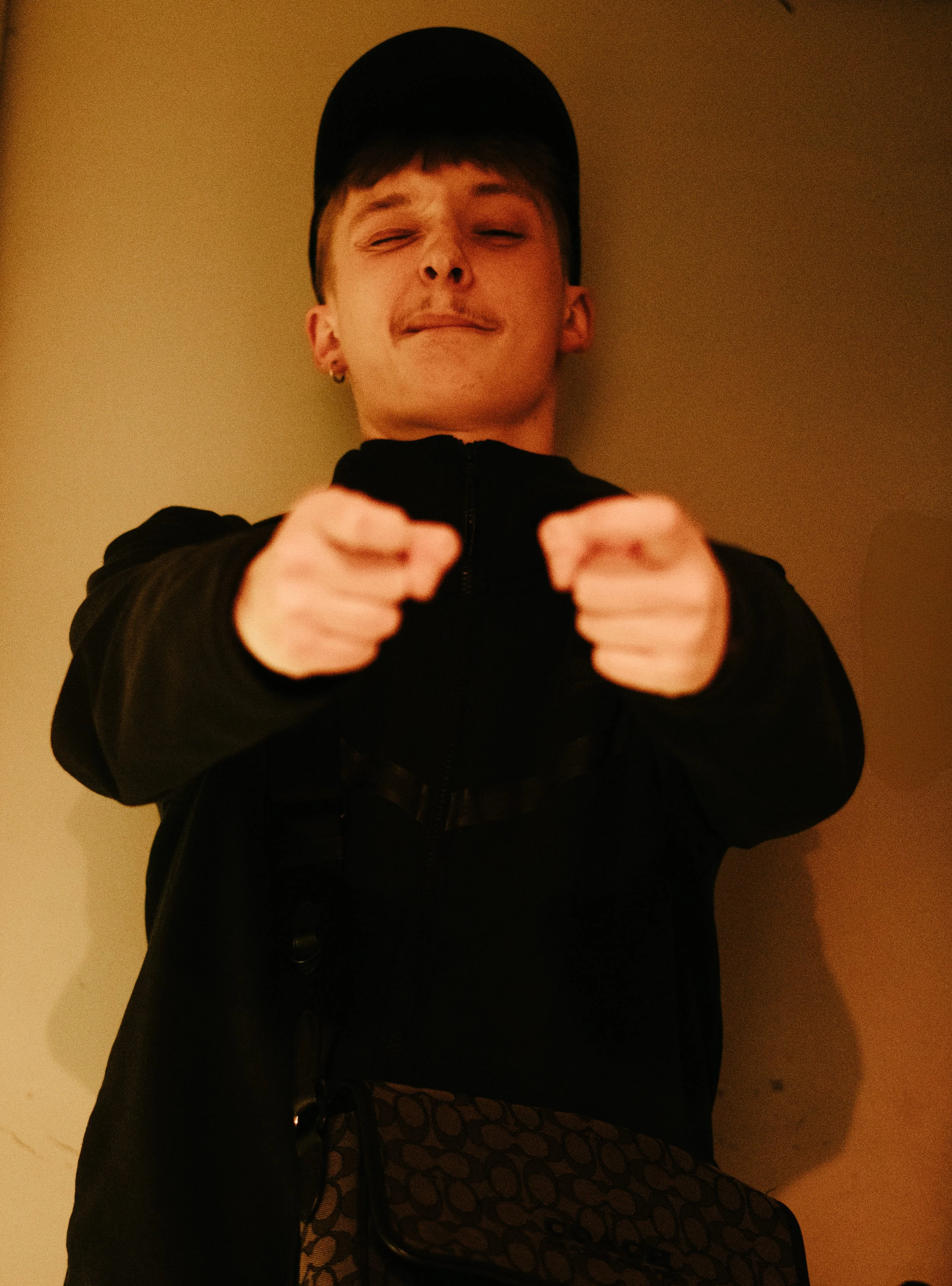 Jeune homme portant un chapeau noir, un sweat noir, et un sac à main avec un motif circulaire, faisant un geste vers la caméra dans un mur beige.