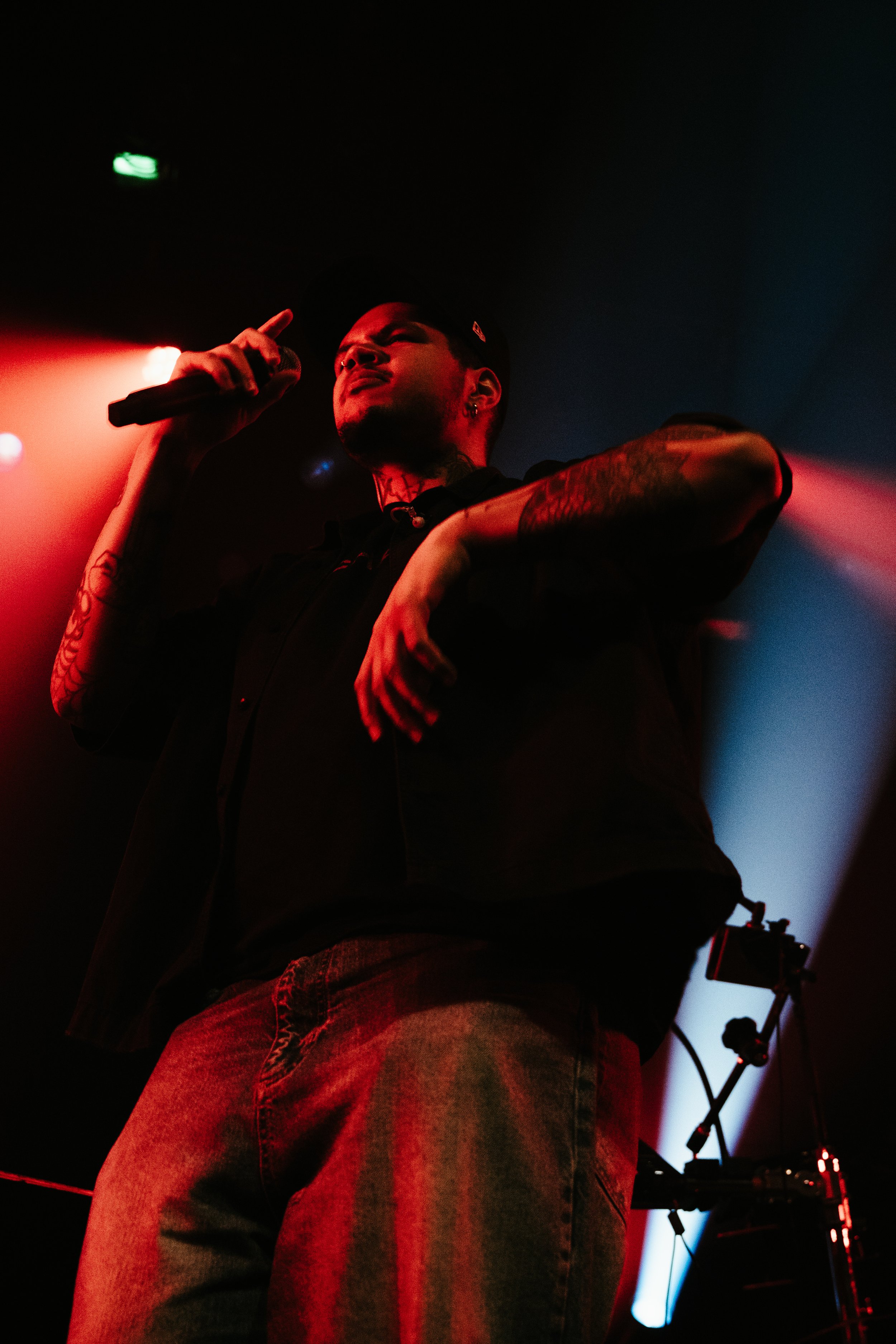 Un jeune homme avec des tatouages, portant une casquette, chante ou parle dans un micro lors d'une performance sur scène avec un éclairage rouge et bleu.