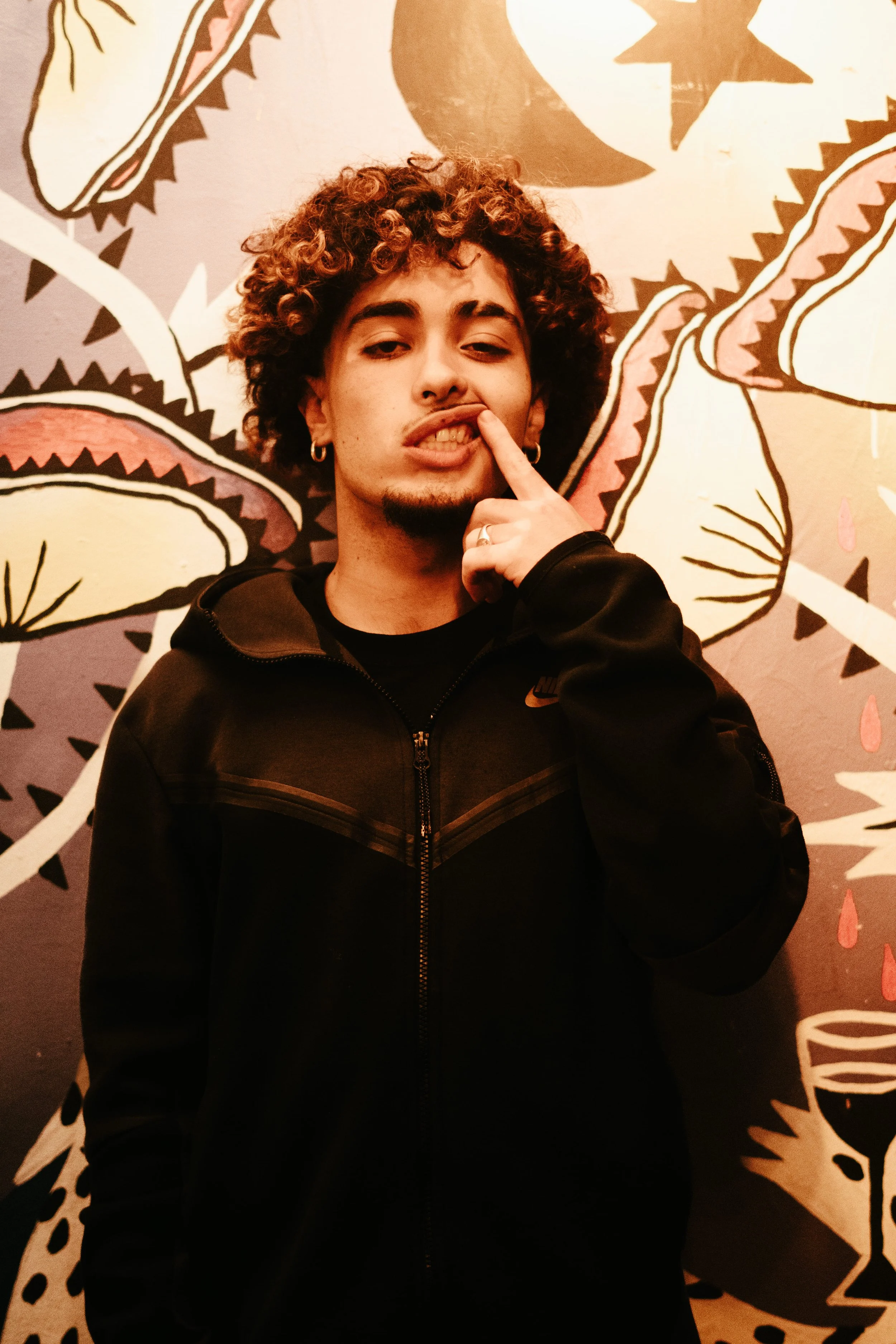 Jeune homme avec des cheveux frisés, portant un sweat noir, posant avec un doigt à la bouche devant un mur décoré avec un motif florale stylisé.