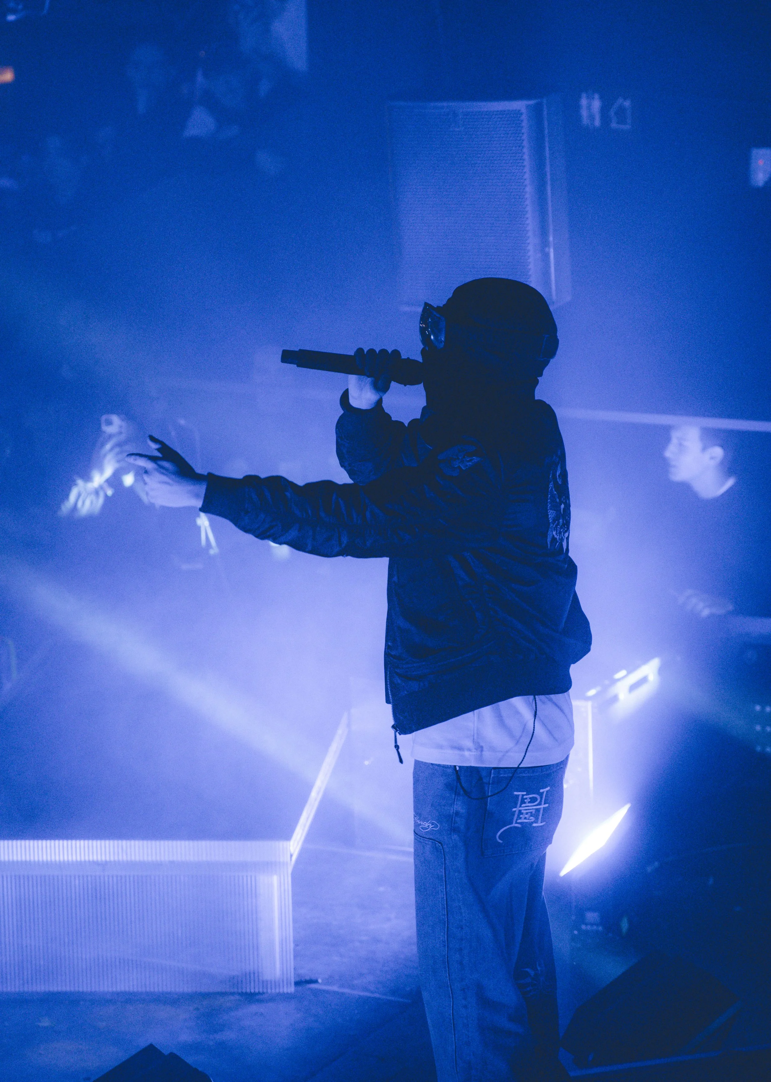 Un artiste de rap en scène lors d'un spectacle, portant une veste noire, des lunettes de protection, tenant un micro, éclairé par des lumières bleues.
