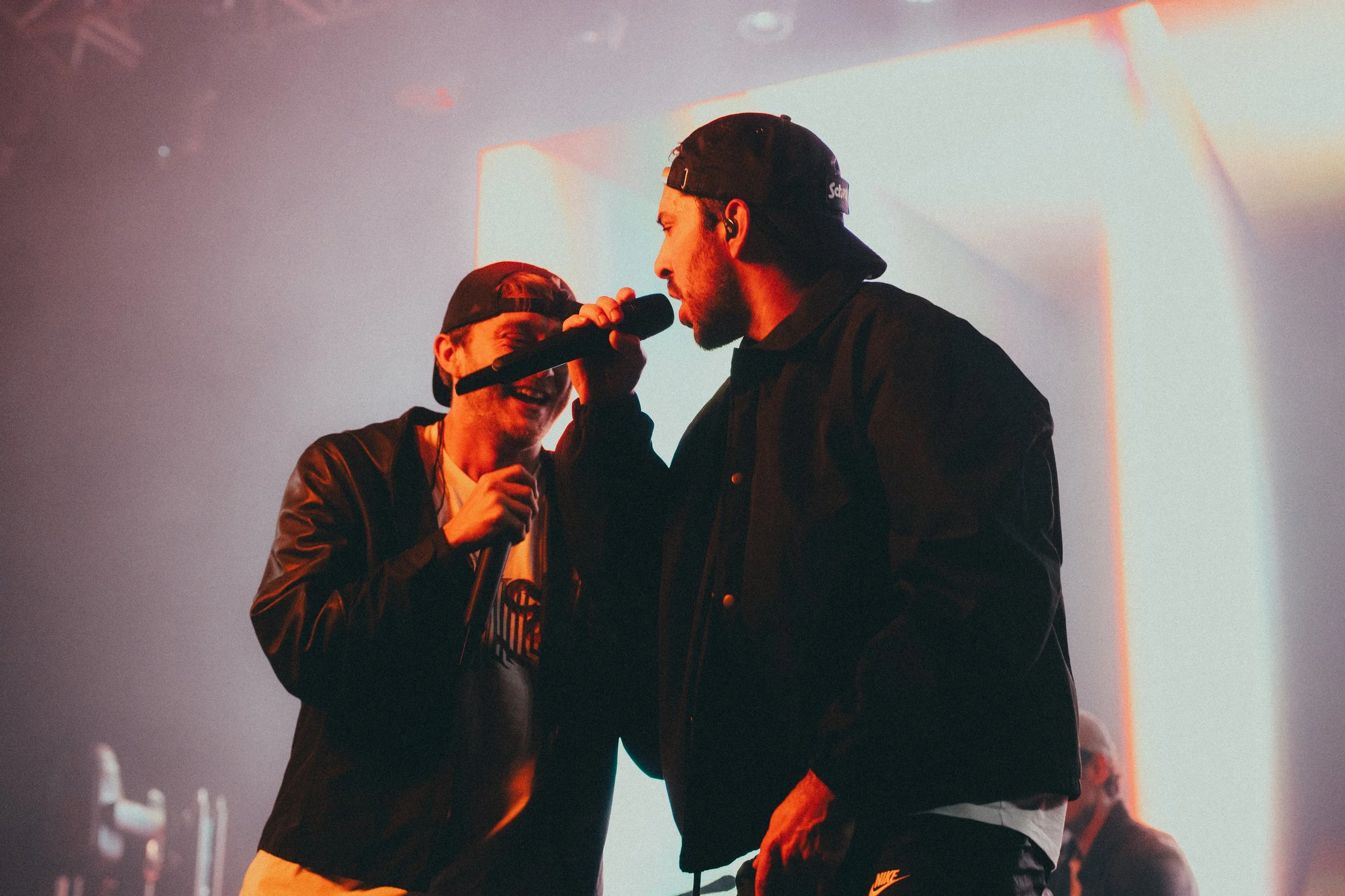 Deux jeunes hommes chantent ensemble sur scène, l'un avec un casque et un micro, l'autre avec un chapeau, lors d'un concert.