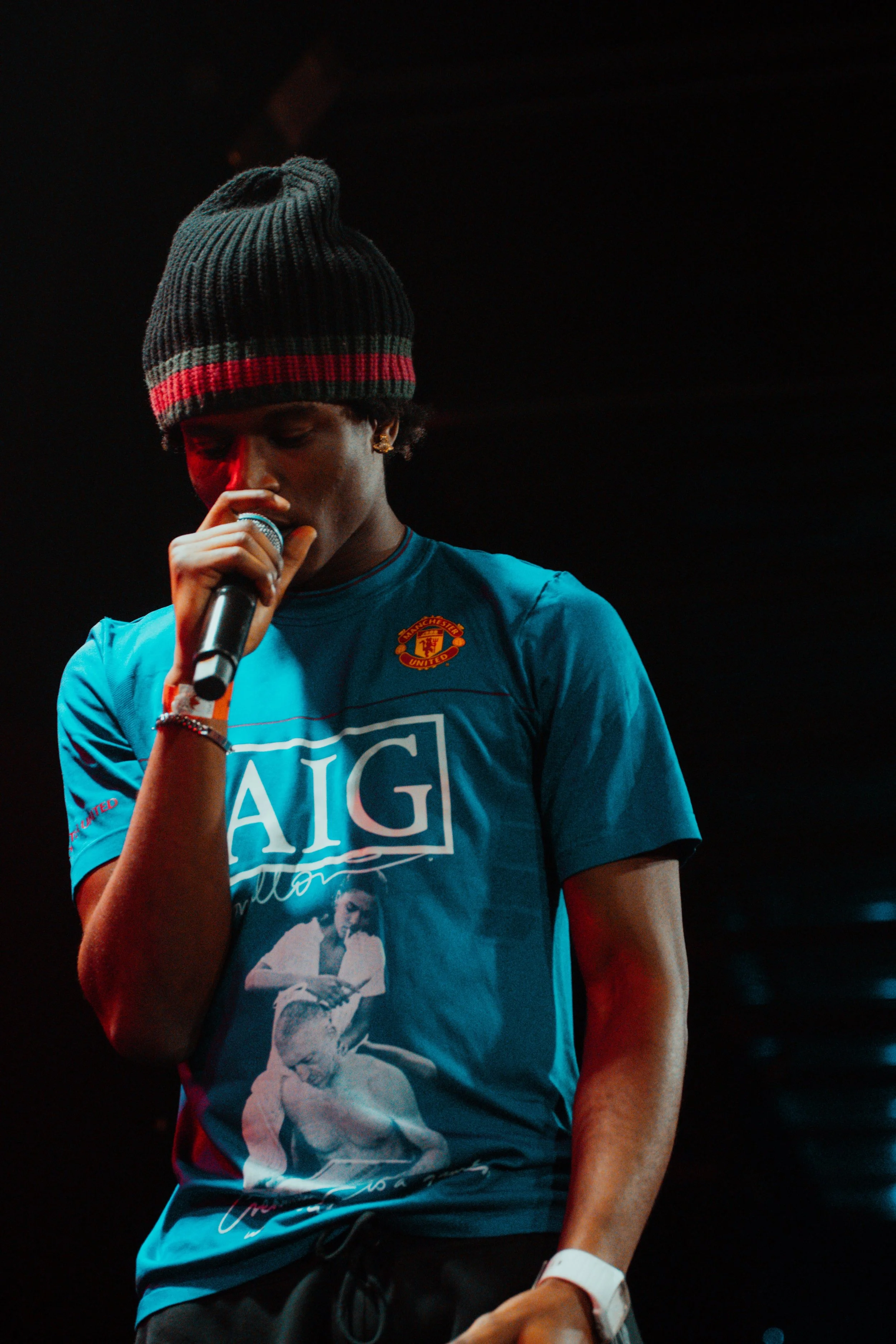 Une personne portant un bonnet noir avec une bande rouge, tenant un microphone, portant un t-shirt bleu avec le logo de Manchester United et une image d'une scène de gladiateurs, sur scène avec un éclairage foncé.