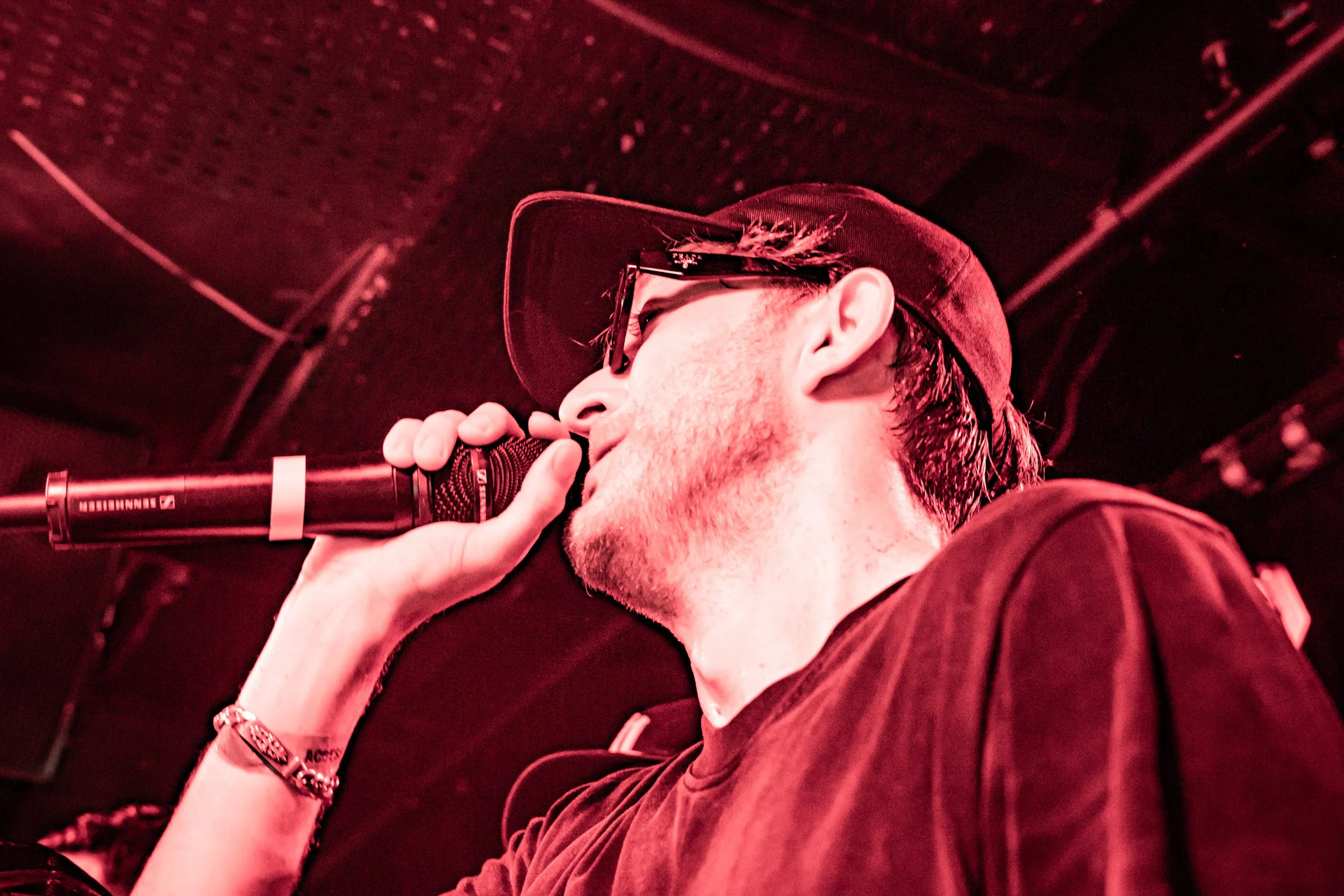 Un homme portant une casquette, des lunettes et un bracelet, chantant ou parlant dans un microphone dans un environnement sombre avec un éclairage rouge.