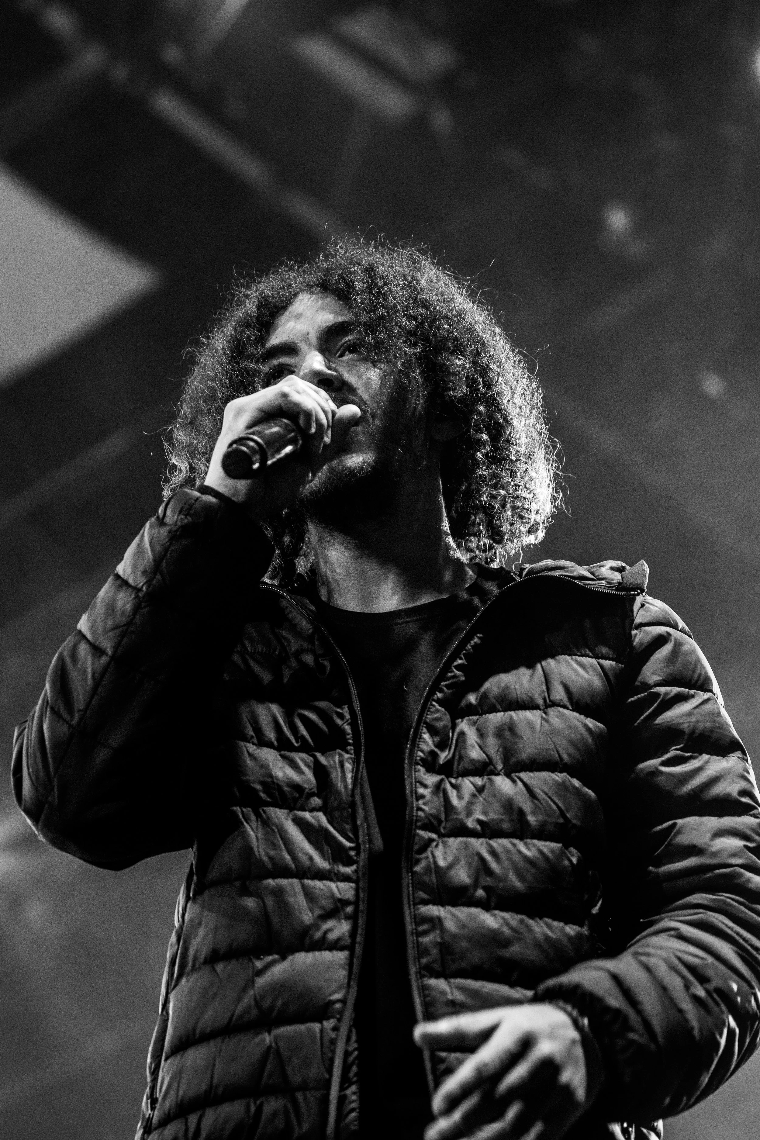 Un homme avec des cheveux bouclés et une barbe tient un microphone et chante ou parle lors d'une performance, en noir et blanc.