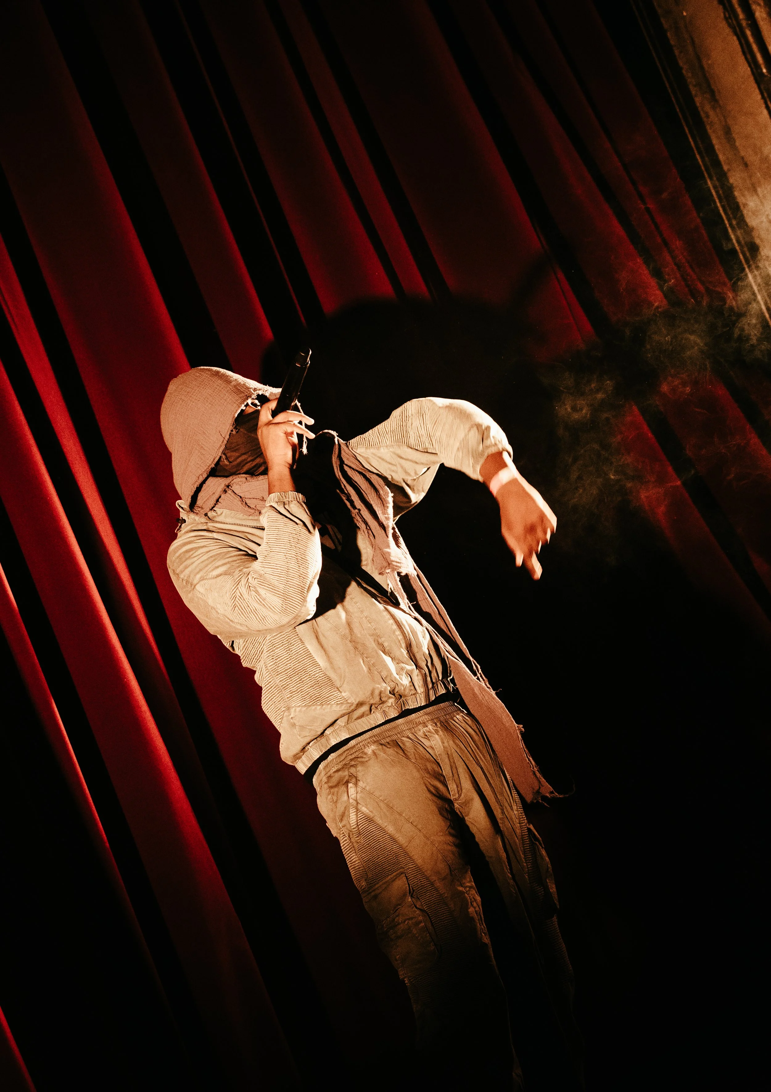 Personne portant une veste beige et un chapeau, fumant une cigarette, sur fond de rideaux rouges.