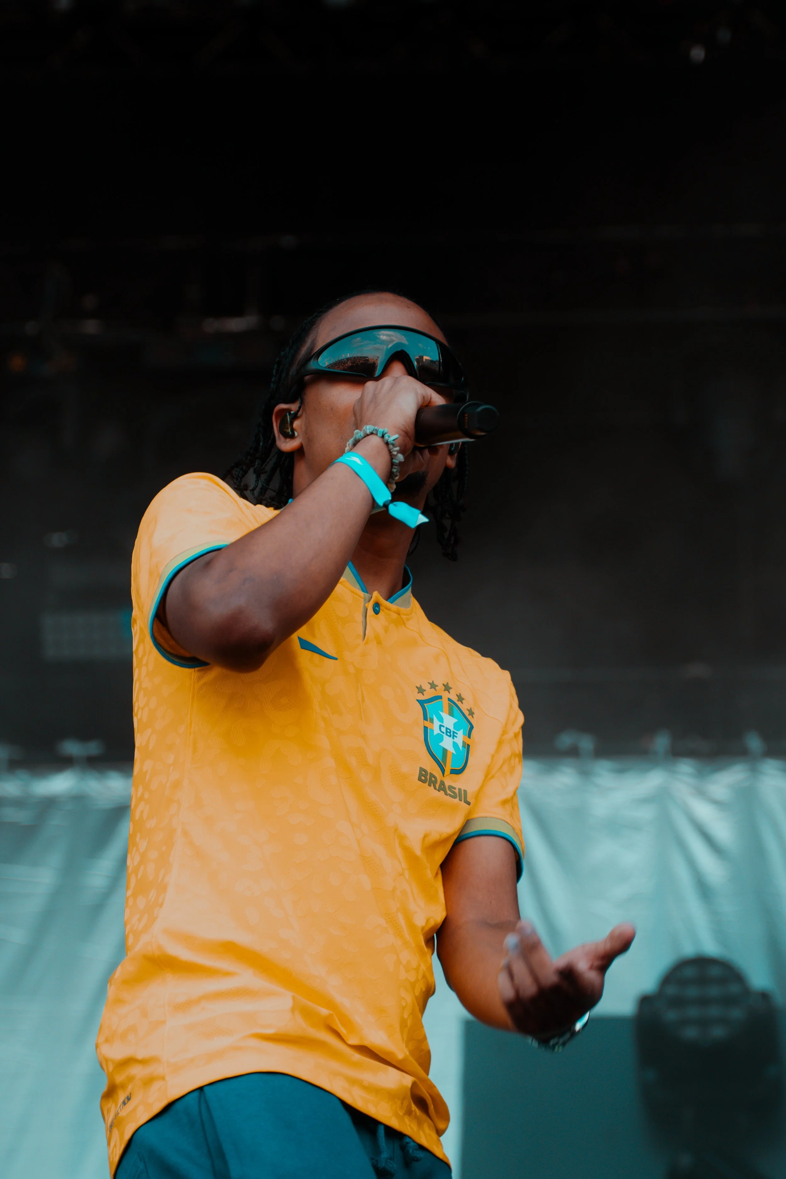 Un chanteur ou un rappeur brésilien portant un maillot de l'équipe nationale de football du Brésil, chantant ou parlant dans un micro lors d'une performance en concert.