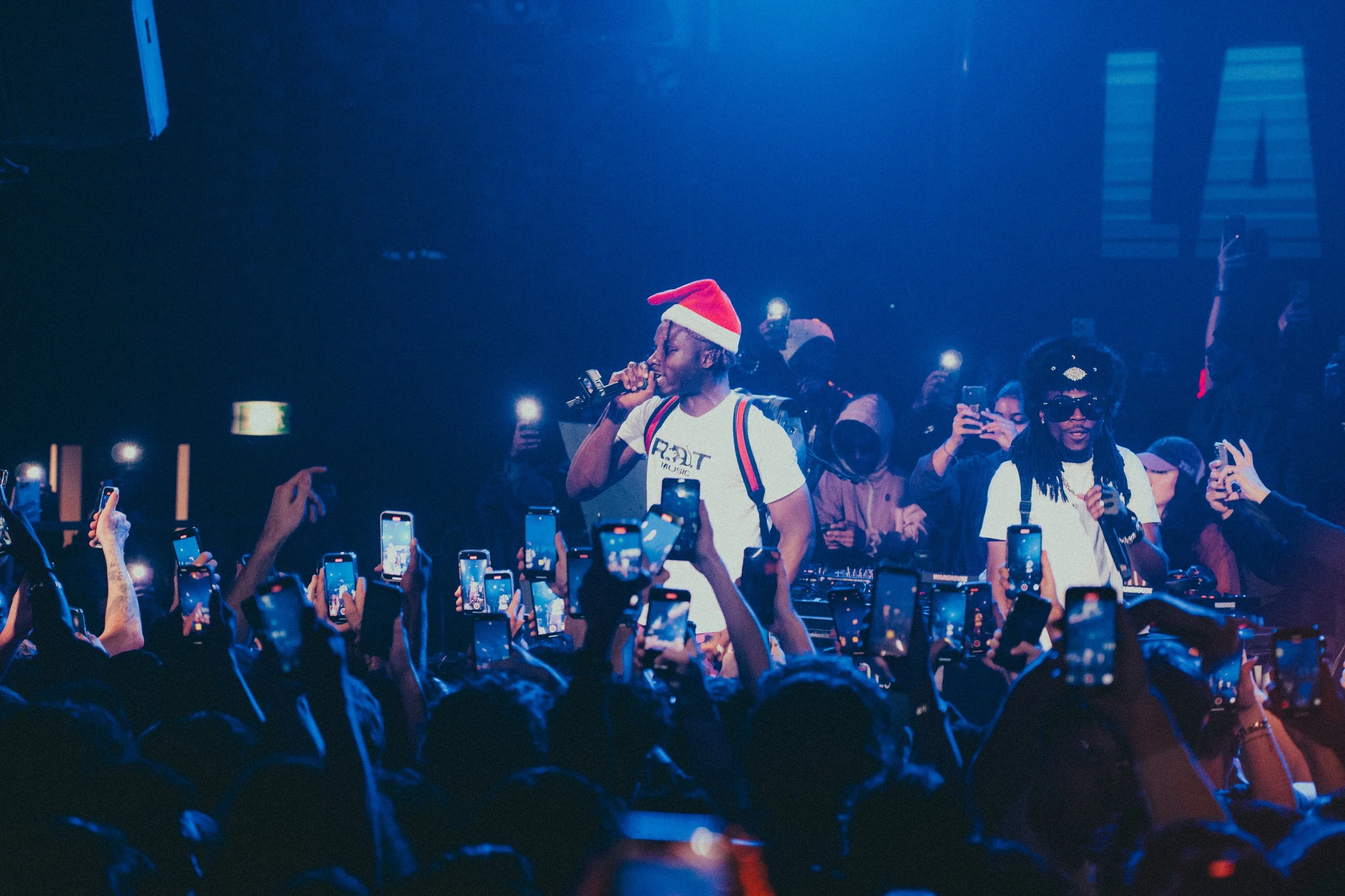 Un concert avec un artiste portant un chapeau de Père Noël, chantant dans un microphone, entouré d'une foule en extase avec des téléphones en l'air, ambiance festive avec lumières bleues.