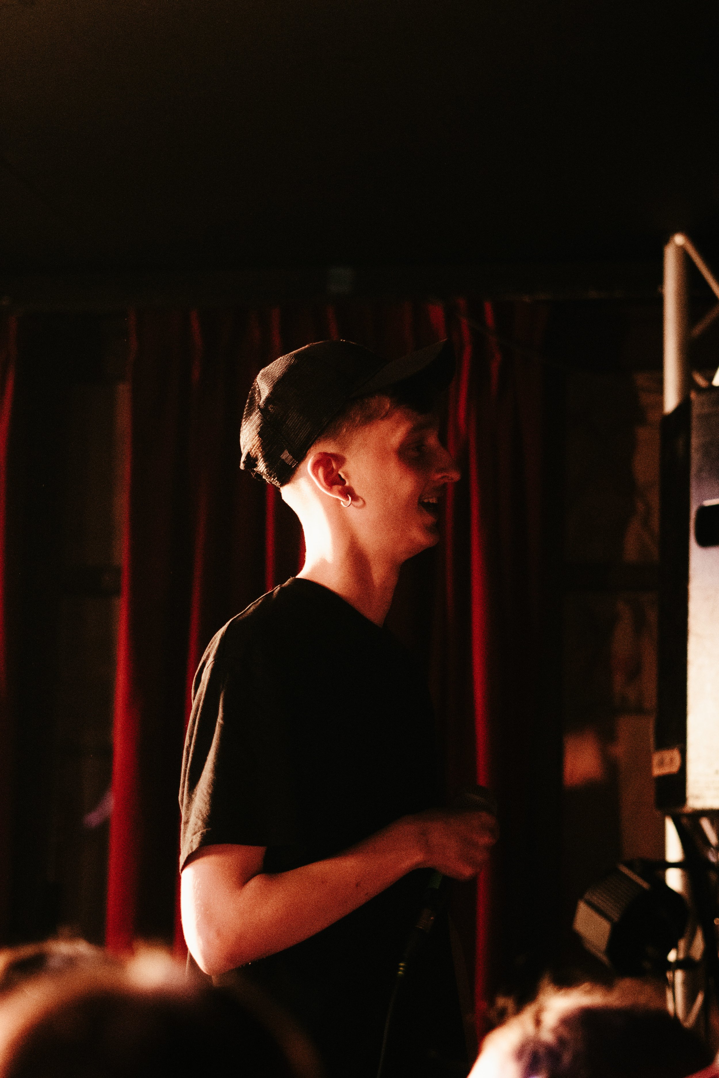 Une jeune personne souriante portant une casquette noire, tenant un microphone, dans un environnement sombre avec des rideaux rouges en arrière-plan.
