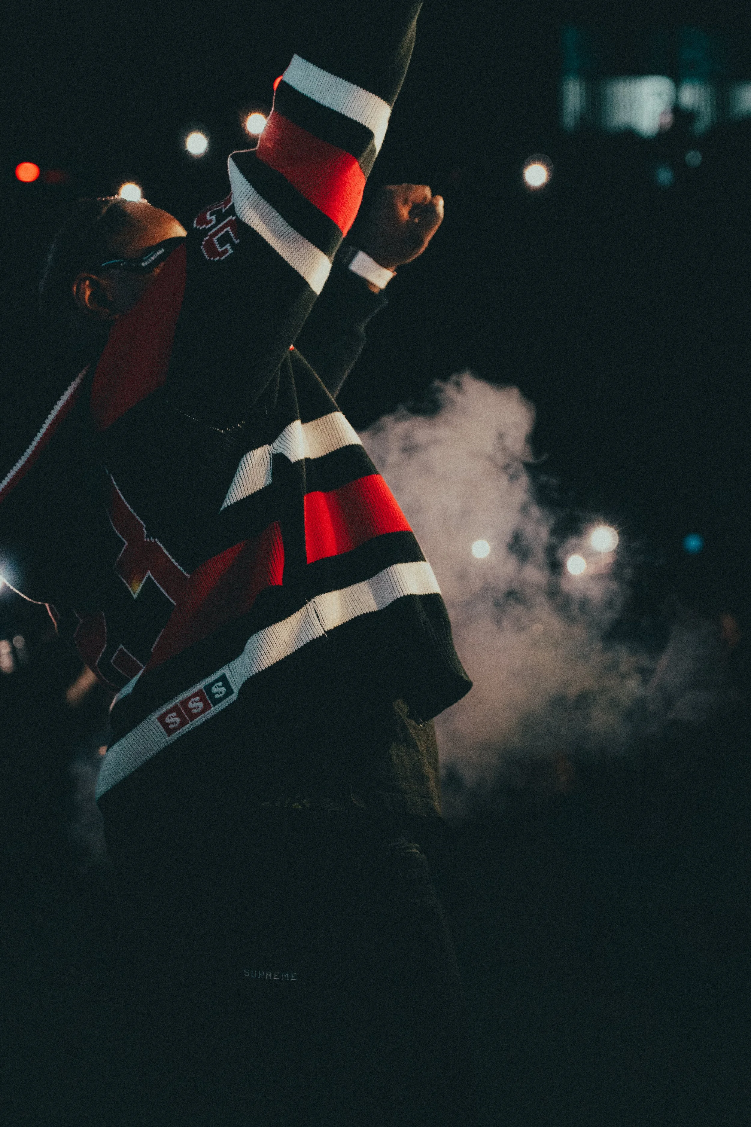 Un homme portant un pull à rayures rouges, blanches et noires lors d'un événement en intérieur, avec de la fumée et des lumières en arrière-plan.