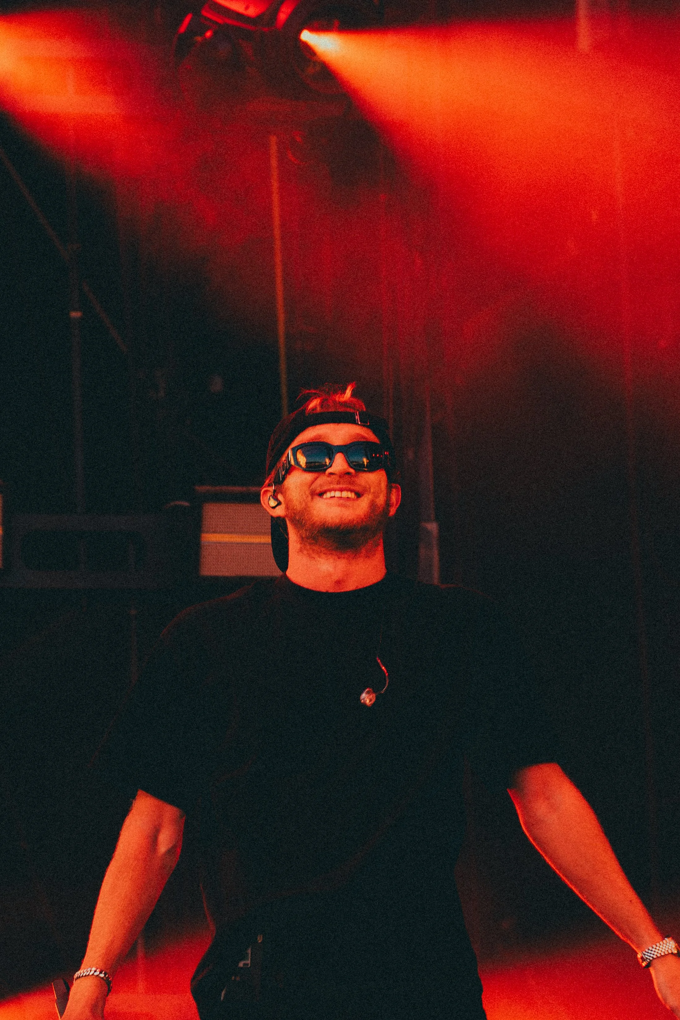 Un homme souriant portant des lunettes de soleil noires, une casquette noire à l'envers et un t-shirt noir, sur une scène de concert avec un éclairage rouge.
