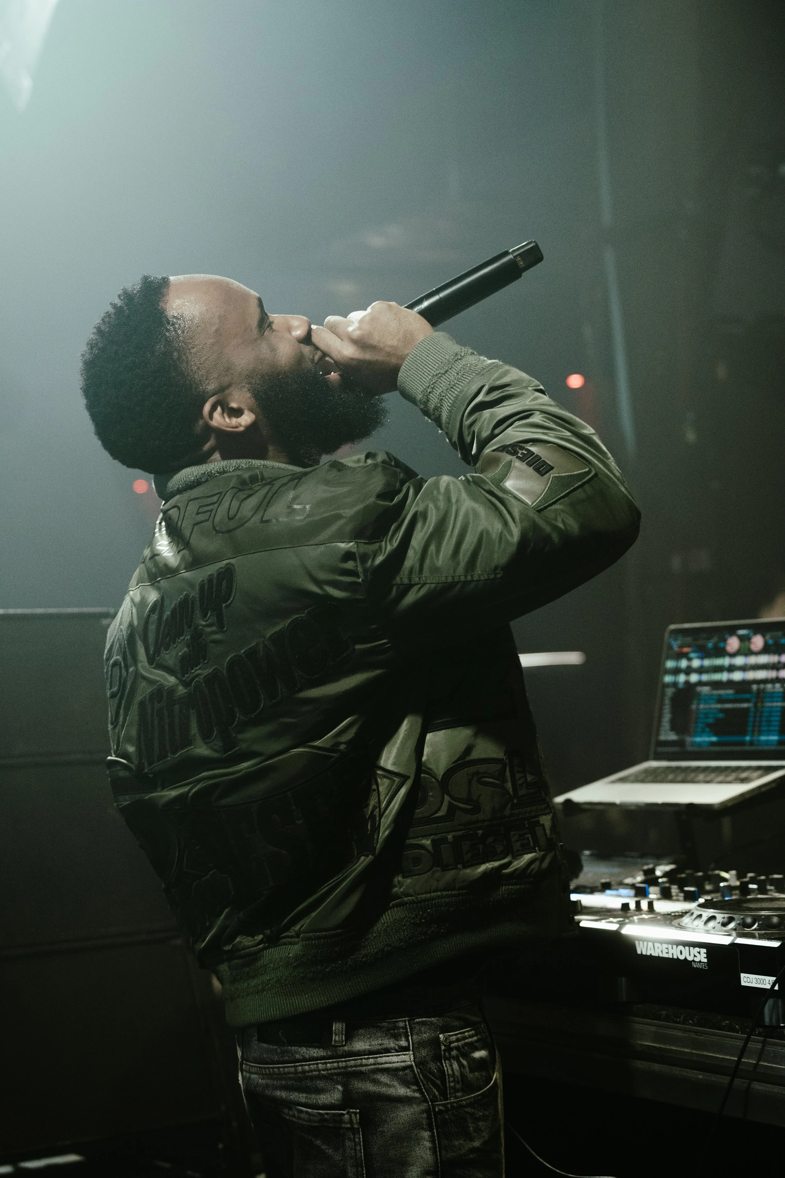 Un homme avec une barbe et des cheveux courts, portant une veste verte, chante en tenant un microphone dans une salle de concert, avec un ordinateur portable et un clavier DJ à proximité.