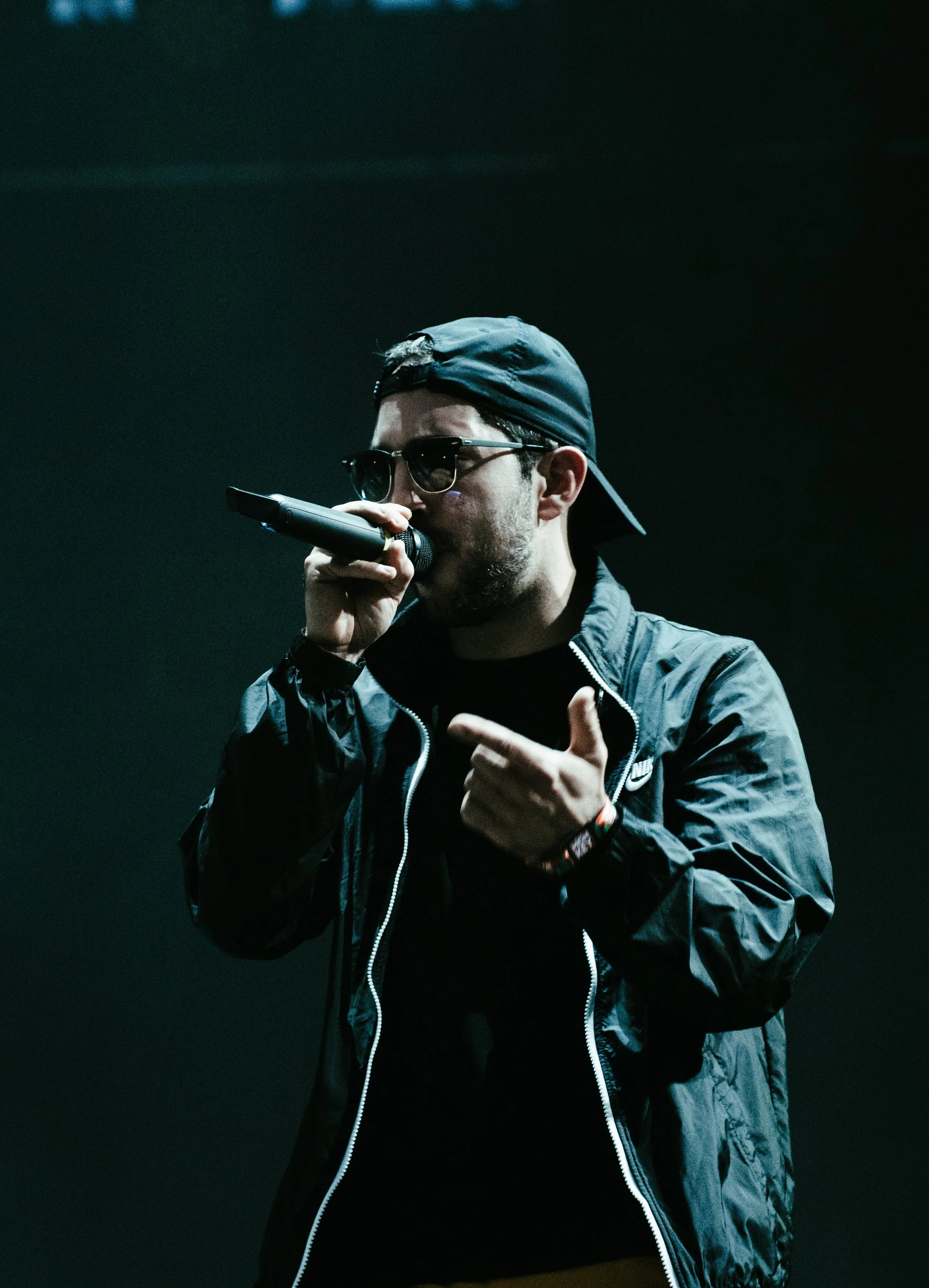 Jeune homme portant une casquette et des lunettes de soleil, tenant un microphone, en train de chanter ou de parler, habillé d'une veste noire dans un environnement sombre.