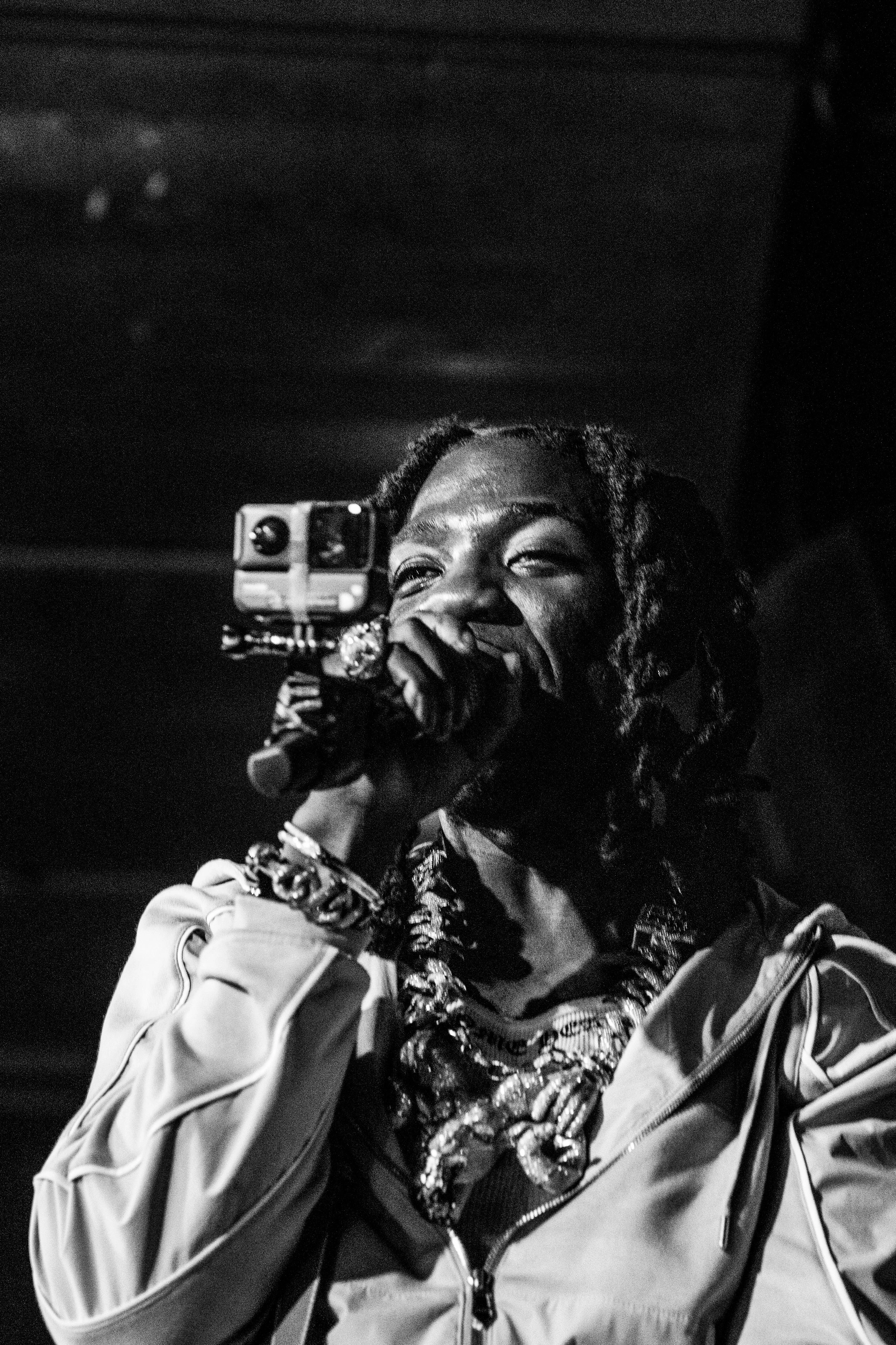Une femme noire portant des bijoux, tenant un microphone et un appareil photo, souriant en chantant ou parlant, habillée d'une veste de sport avec un fond sombre.
