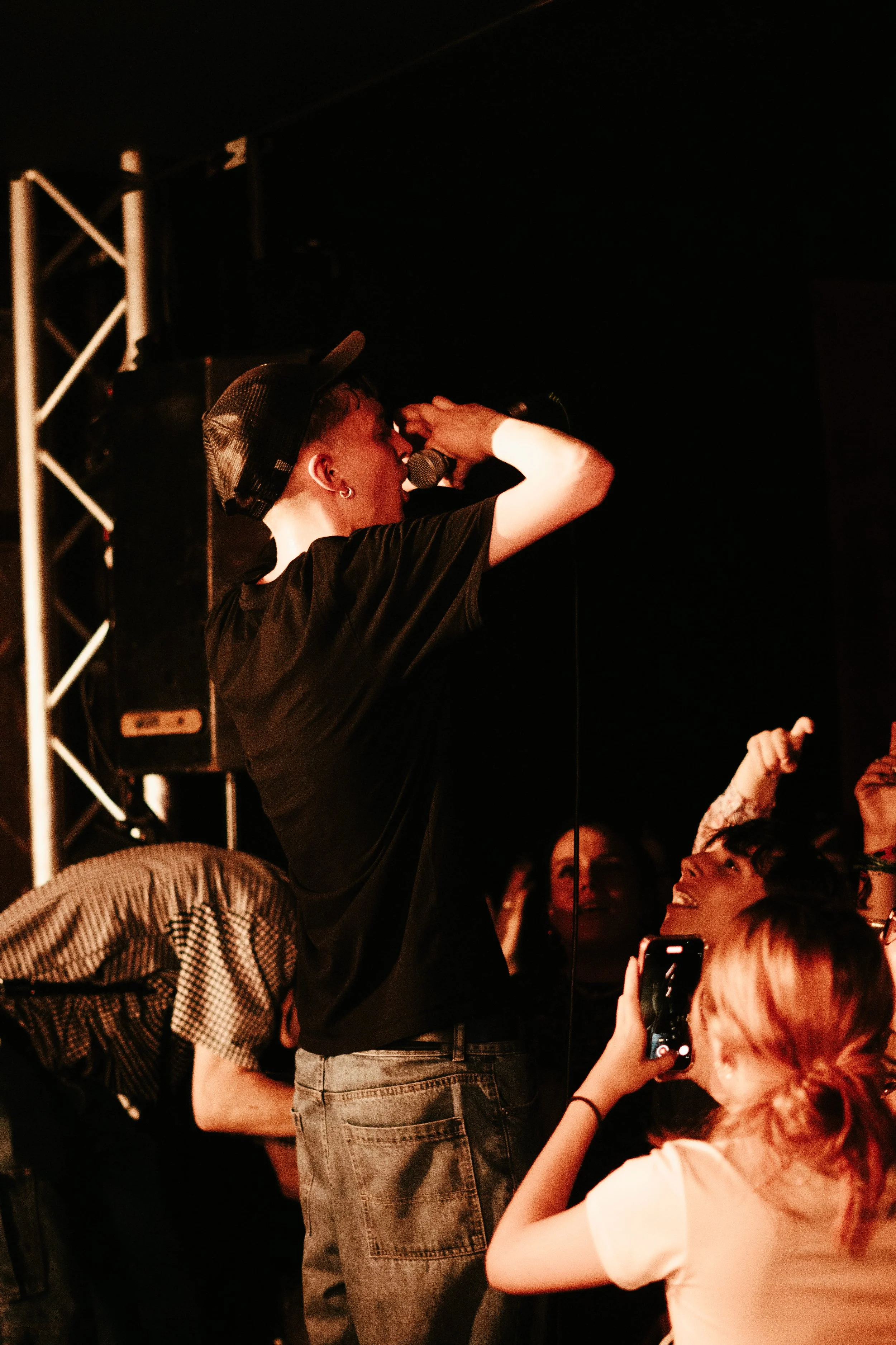 Un jeune homme avec un chapeau de baseball noir, tenant un microphone, en train de chanter ou parler lors d'une performance en concert, entouré d'une audience enthousiaste. La scène est sombre avec un éclairage chaud.