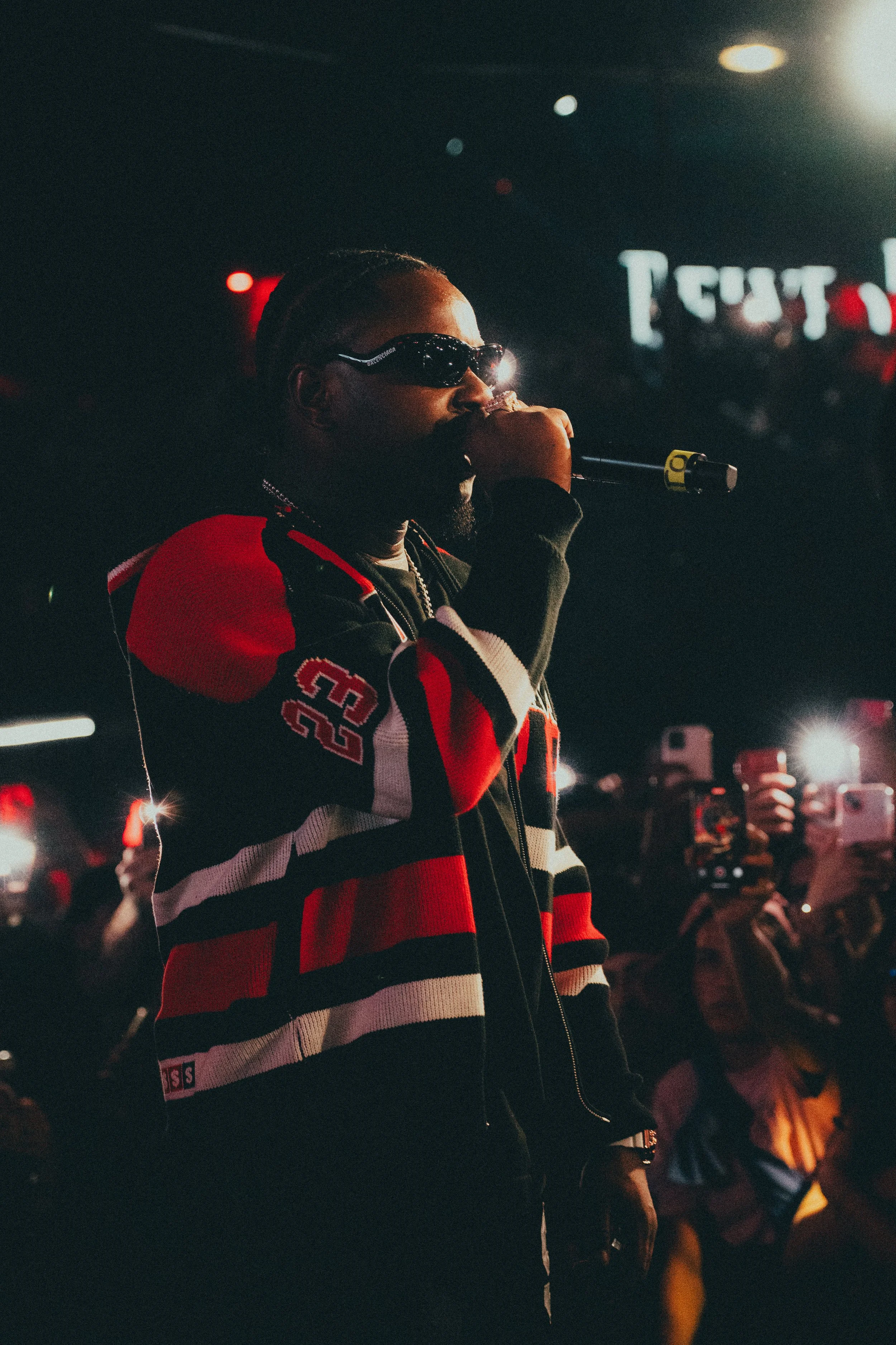 Un homme portant des lunettes de soleil et un sweat à capuche à rayures rouges, noires et blanches, tenant un microphone dans un environnement sombre avec des lumières et des appareils photo en arrière-plan.