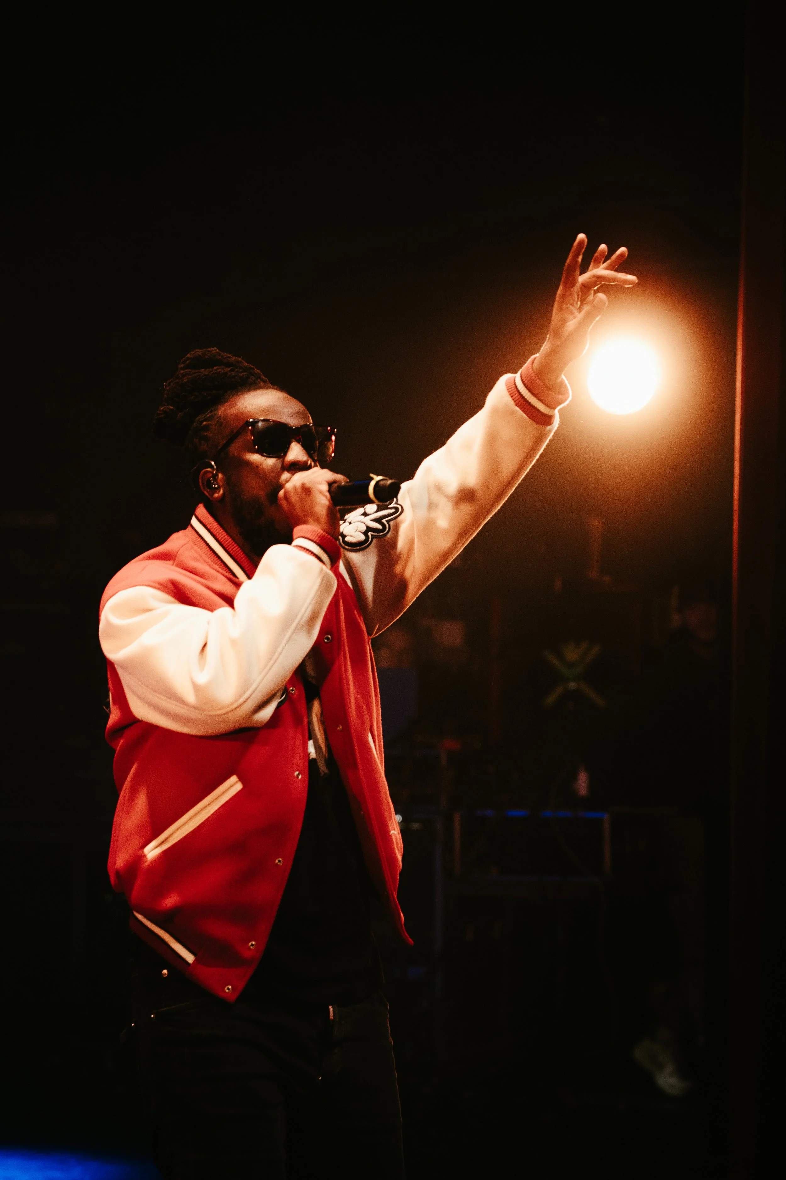 Un homme portant des lunettes de soleil, un sweat à capuche et une veste rouge, chante dans un microphone lors d'une performance sur scène, avec un éclairage chaud derrière lui.
