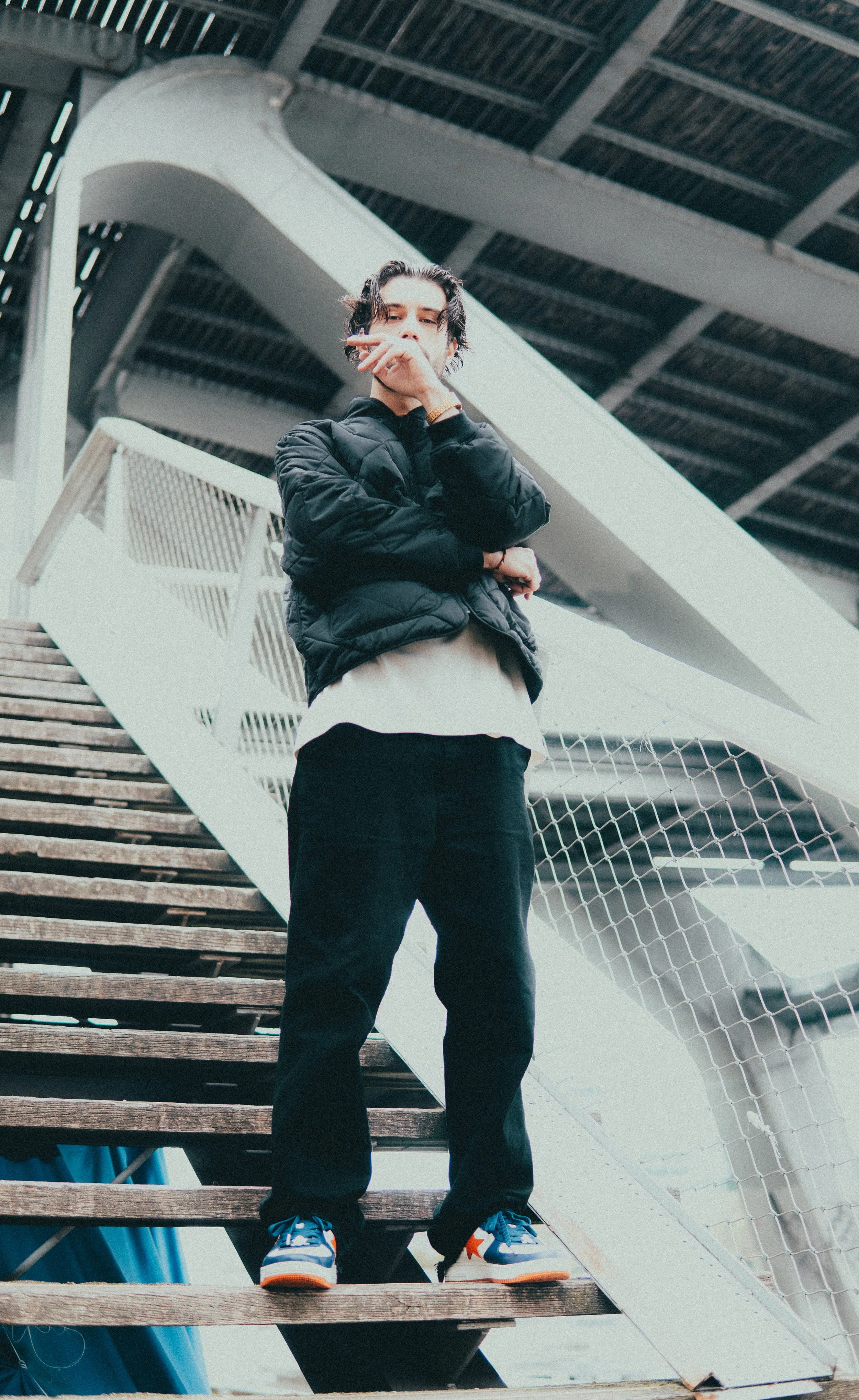 Jeune homme debout sur une échelle de bois, portant une veste noire, un pantalon noir, et des chaussures colorées, sous une structure métallique.