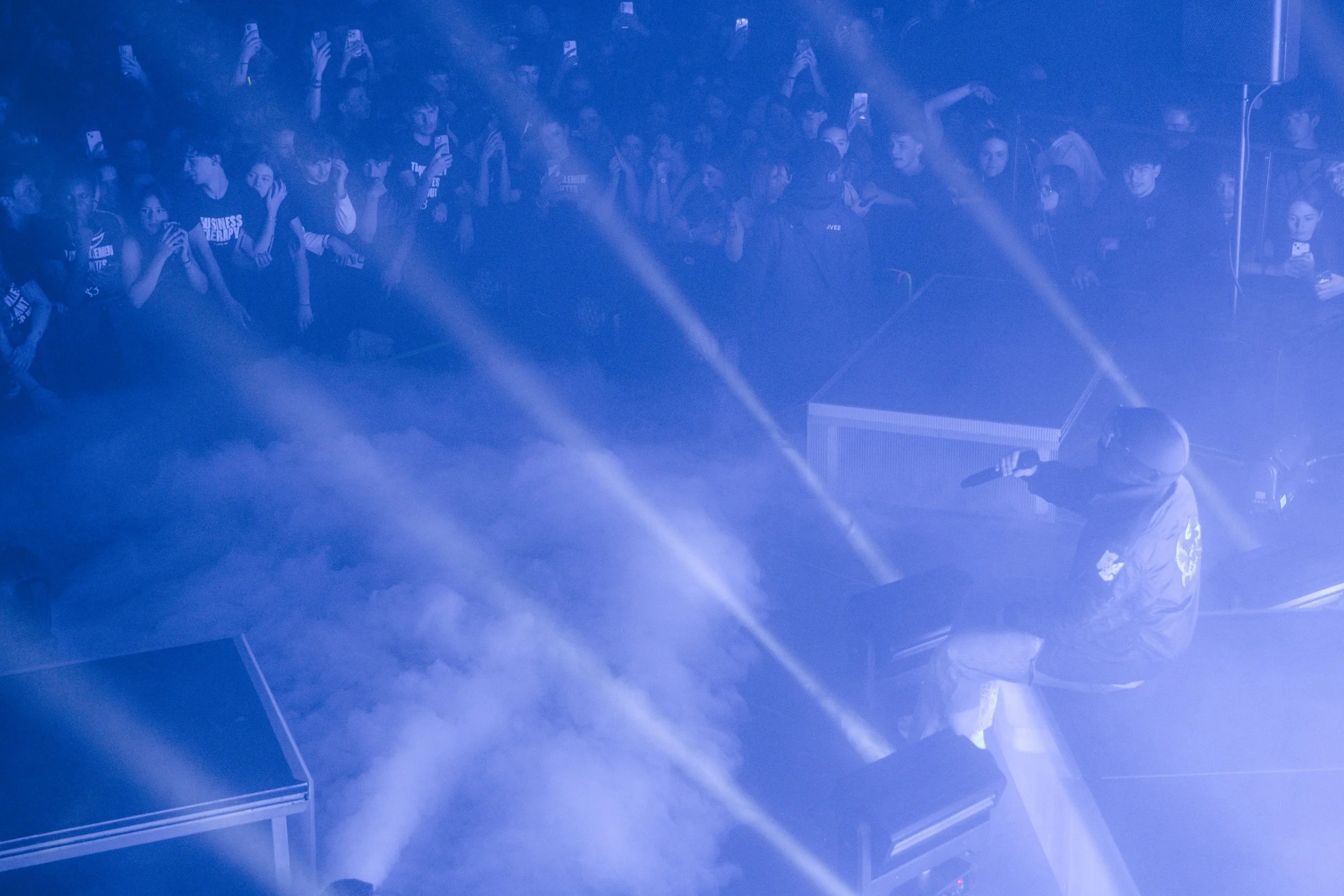 Une scène d'un concert ou d'une performance avec un artiste sur scène entouré de lumières bleues, face à un public nombreux qui regarde et prend des photos.