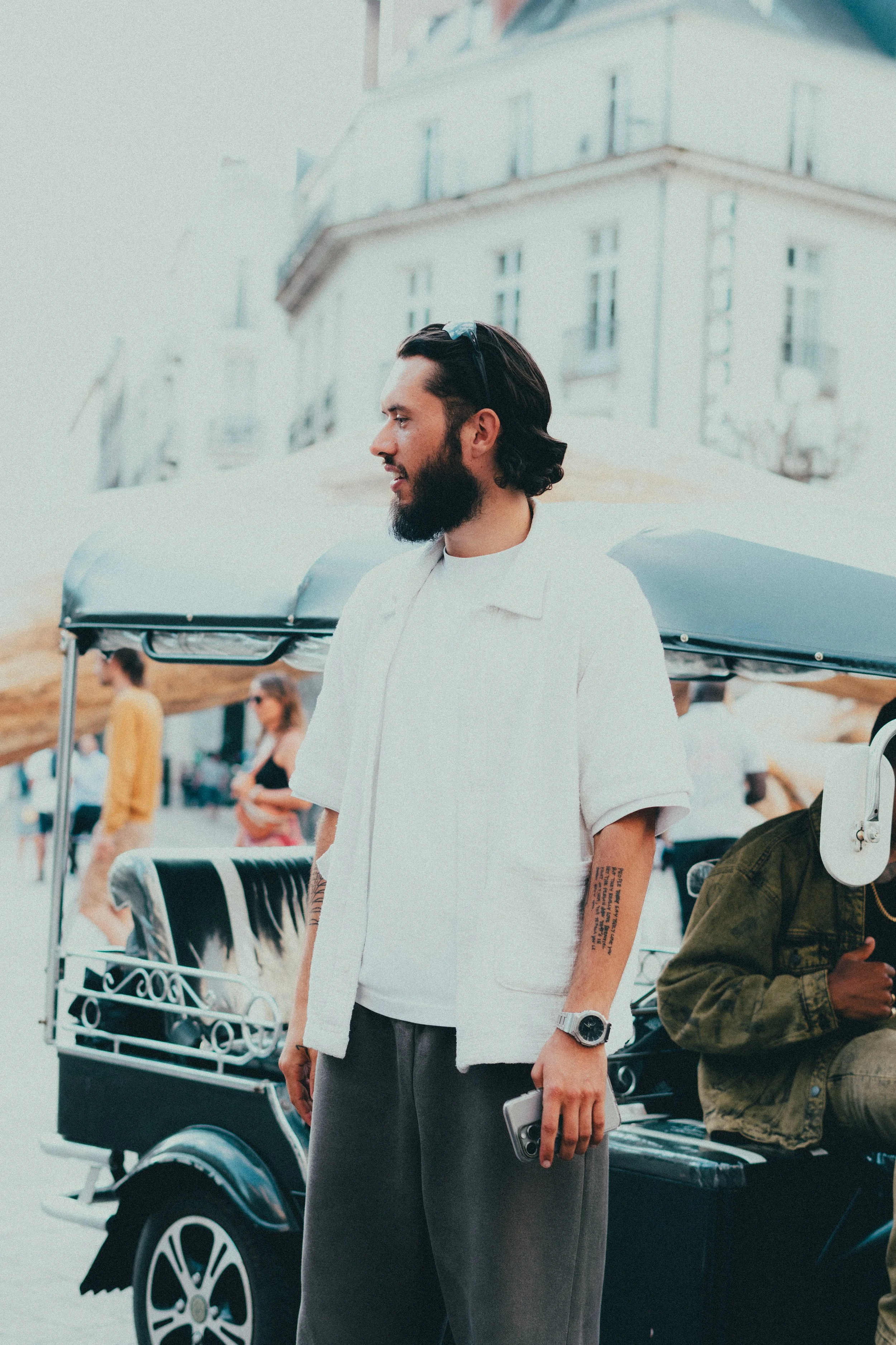 Homme avec barbe et lunettes de soleil, portant une chemise blanche, tenant un téléphone, debout devant une voiturette électrique, en arrière-plan des bâtiments et des personnes.