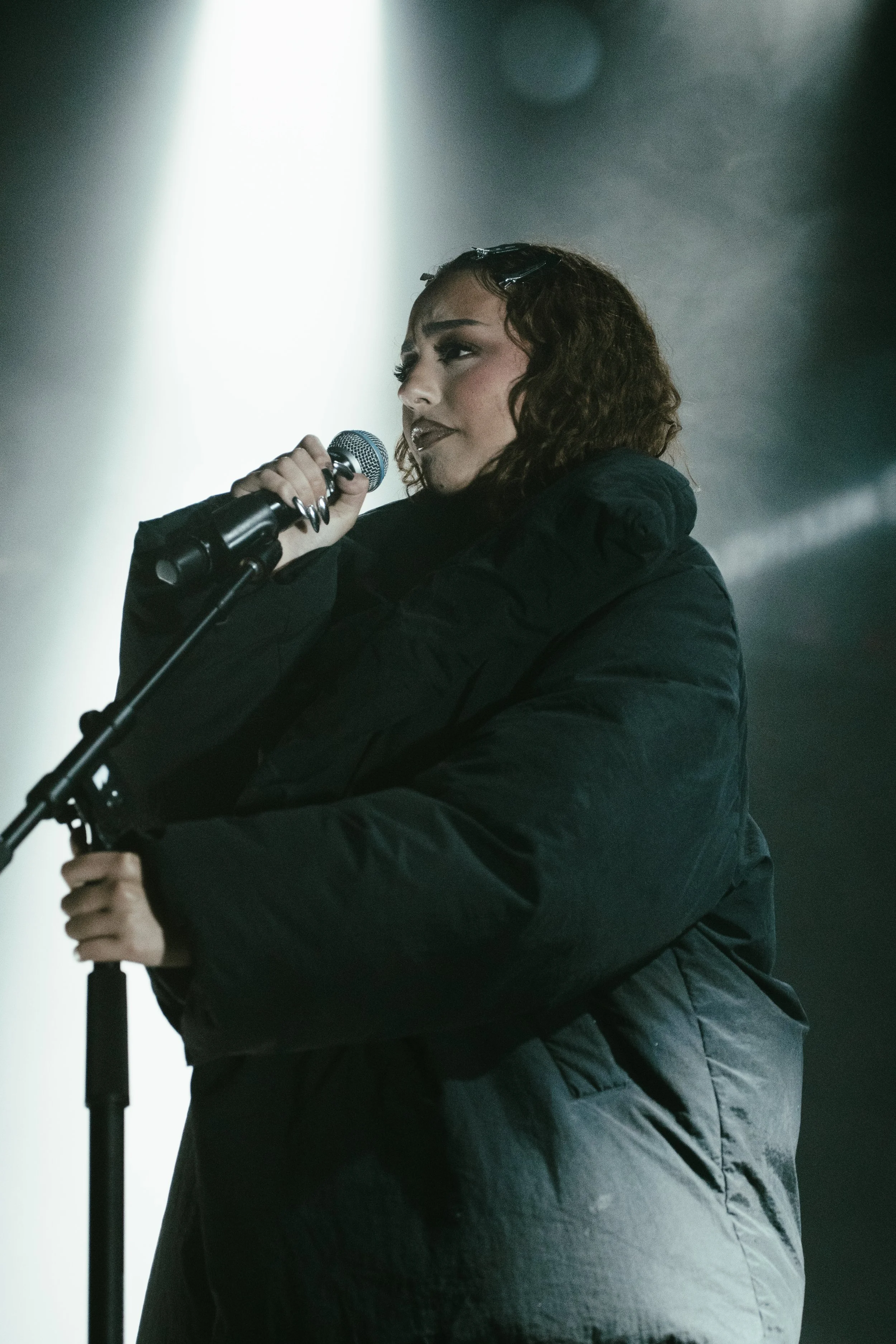 Une femme avec des cheveux bouclés et roux, vêtue d'une veste noire, chante ou parle dans un microphone sur scène, sous un éclairage dramatique.