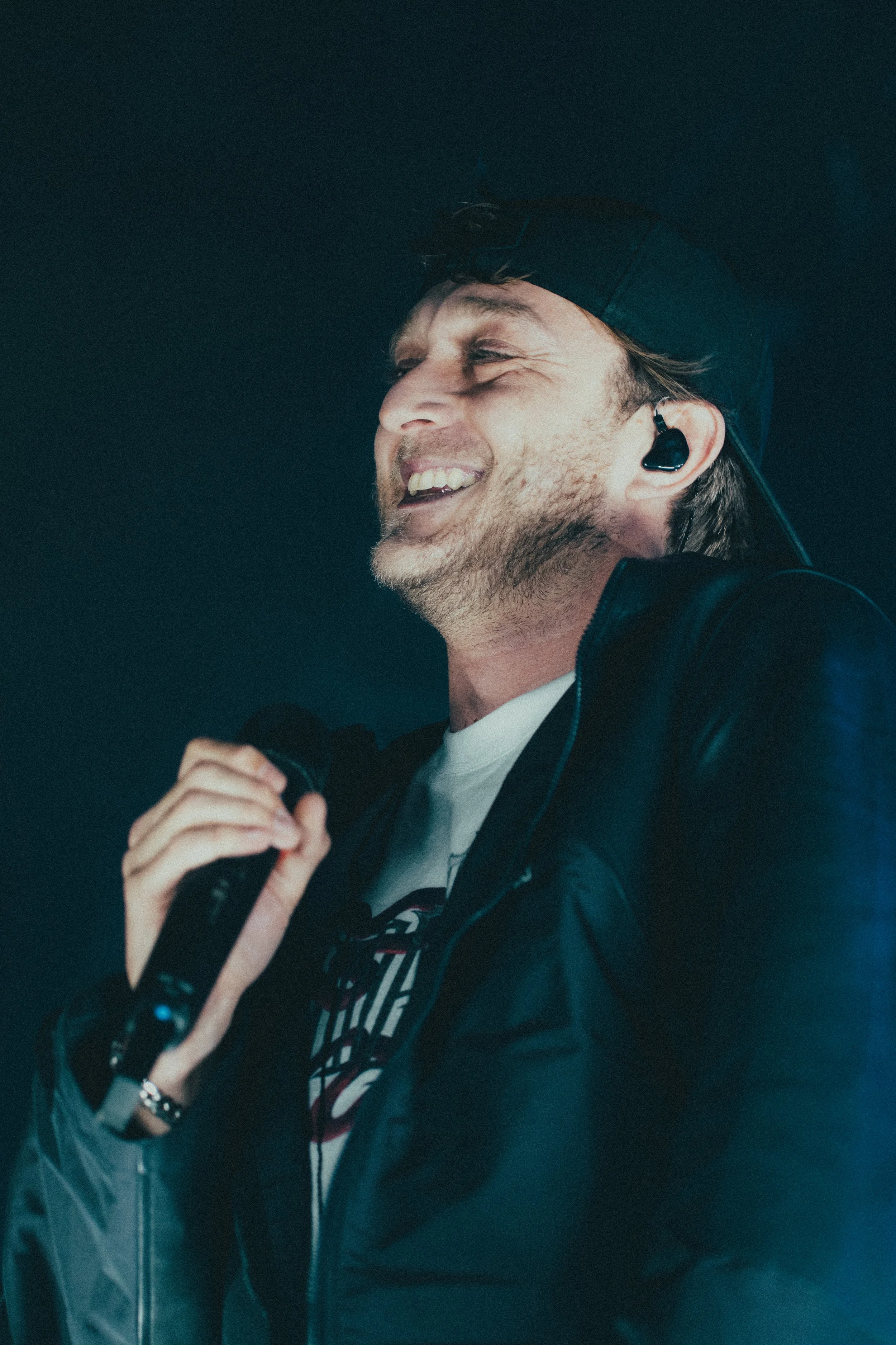 Un homme souriant, portant une casquette et une veste en cuir, tenant un microphone, avec un écouteur sans fil, en train de chanter ou parler.