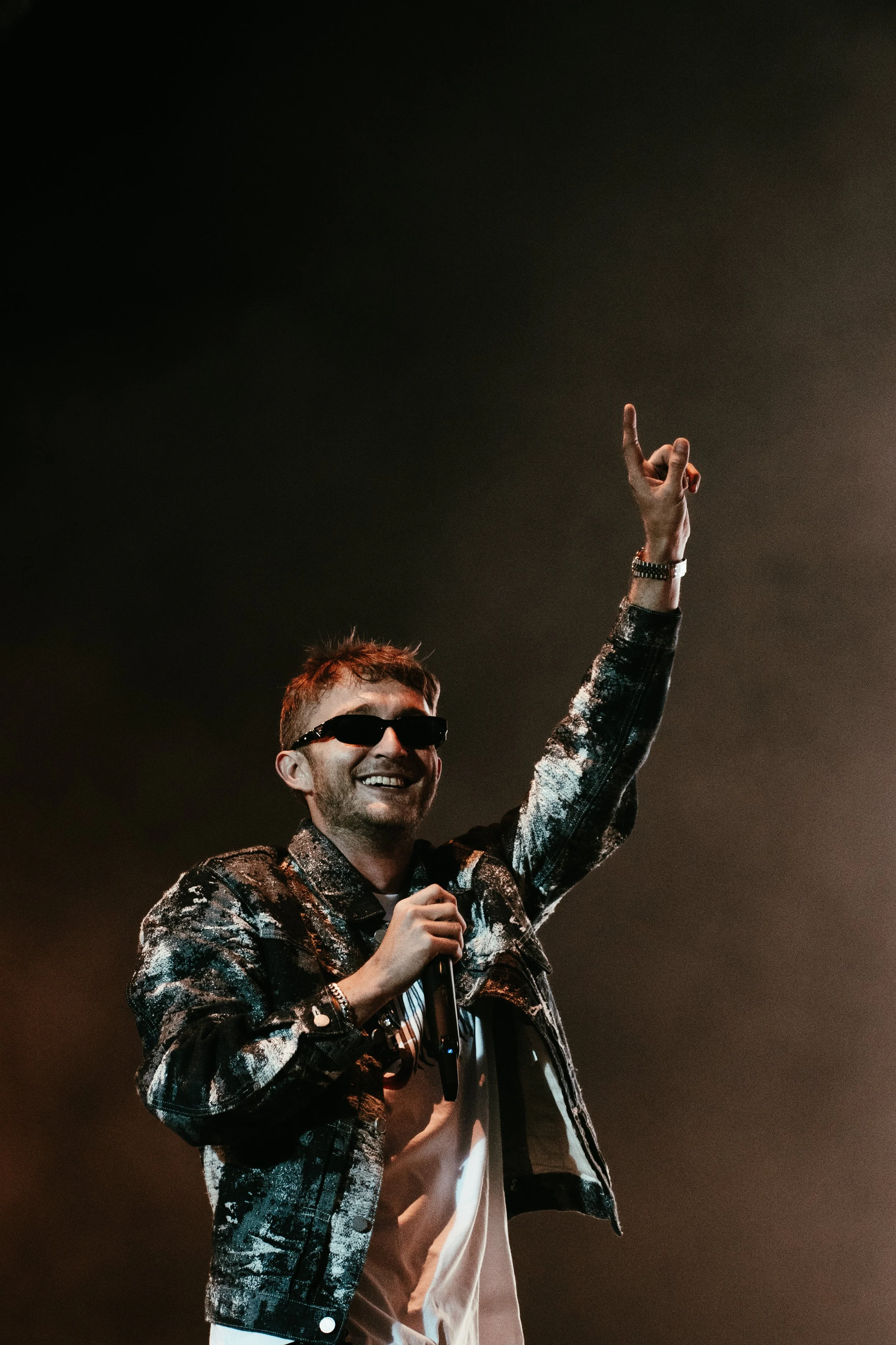 Un homme souriant portant des lunettes de soleil, une veste à paillettes noires et blanches, tenant un microphone, faisant un geste de la main lors d'une performance ou d'un concert sur scène.