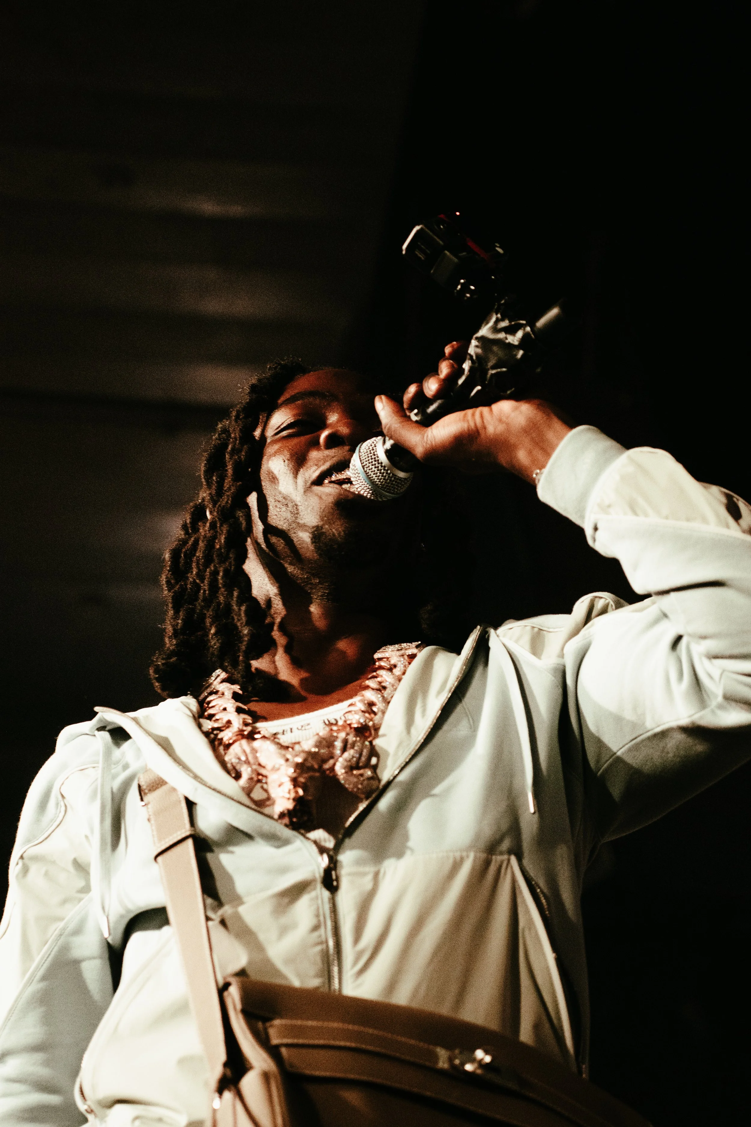 Un homme avec des dreadlocks chantant ou parlant dans un microphone, portant un colliers et un vêtement léger de couleur claire, dans un environnement sombre.