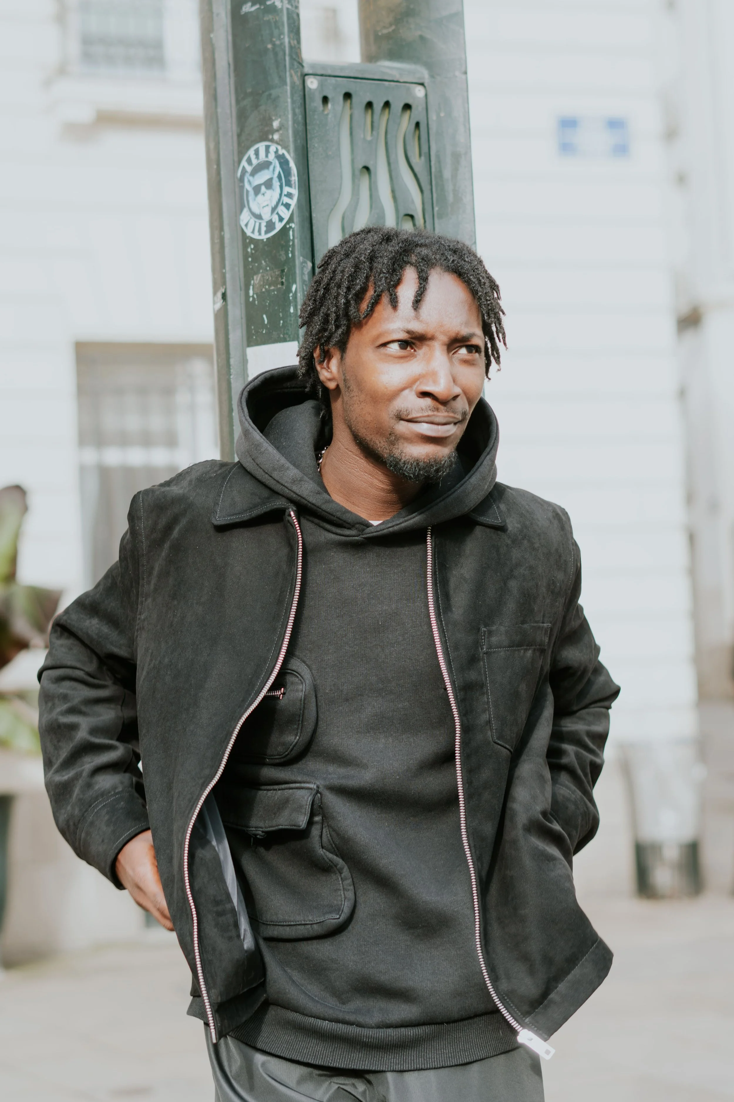 Un homme debout près d'une boîte aux lettres ou d'un panneau électrique, portant un sweat à capuche et une veste noire, avec une expression perplexe ou pensif.