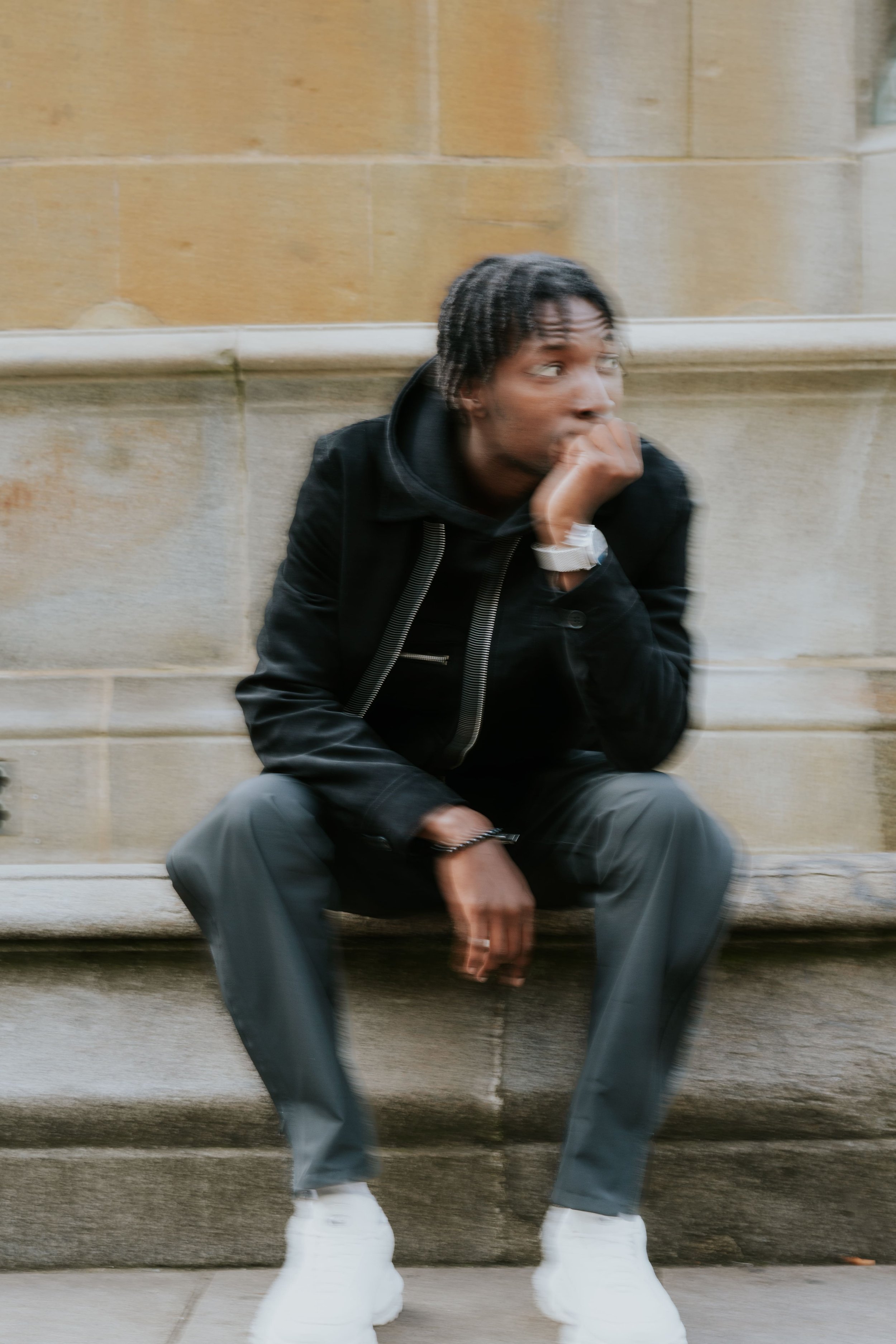 Jeune homme assis sur un banc en pierre, portant une veste noire, pantalon gris, et des baskets blanches, avec la main devant le visage, dans un environnement urbain.