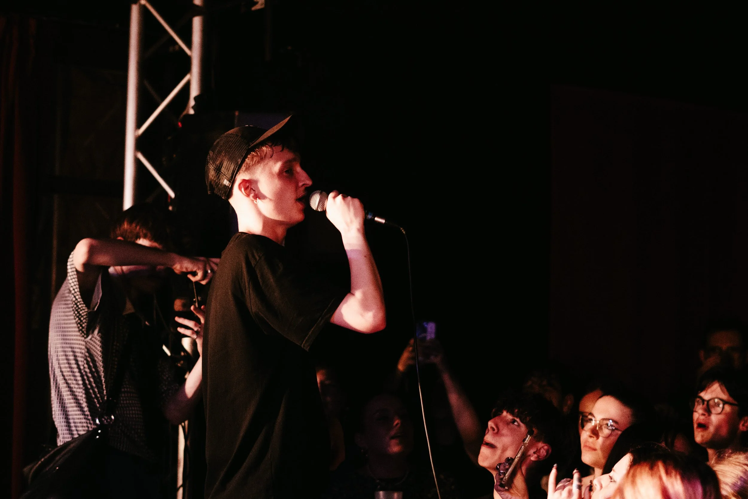 Une jeune femme avec une casquette noire chantant dans un micro lors d'une performance, entourée par un public enthousiaste.