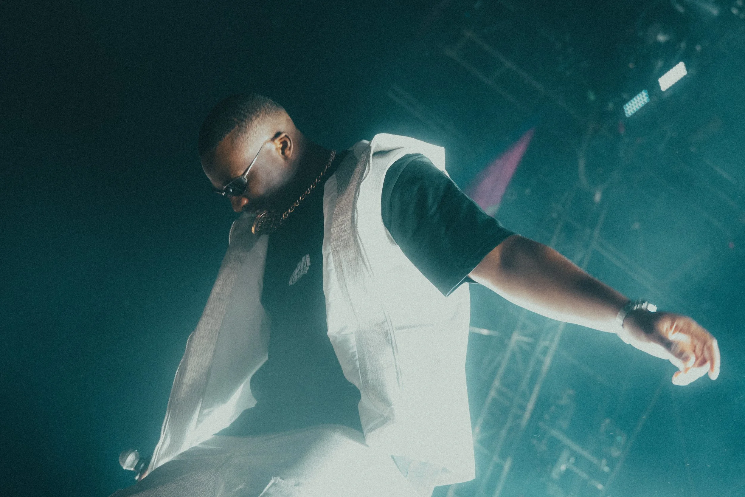 Un artiste de musique en concert, portant des lunettes de soleil, une veste blanche et un t-shirt noir, avec un éclairage de scène dramatique.