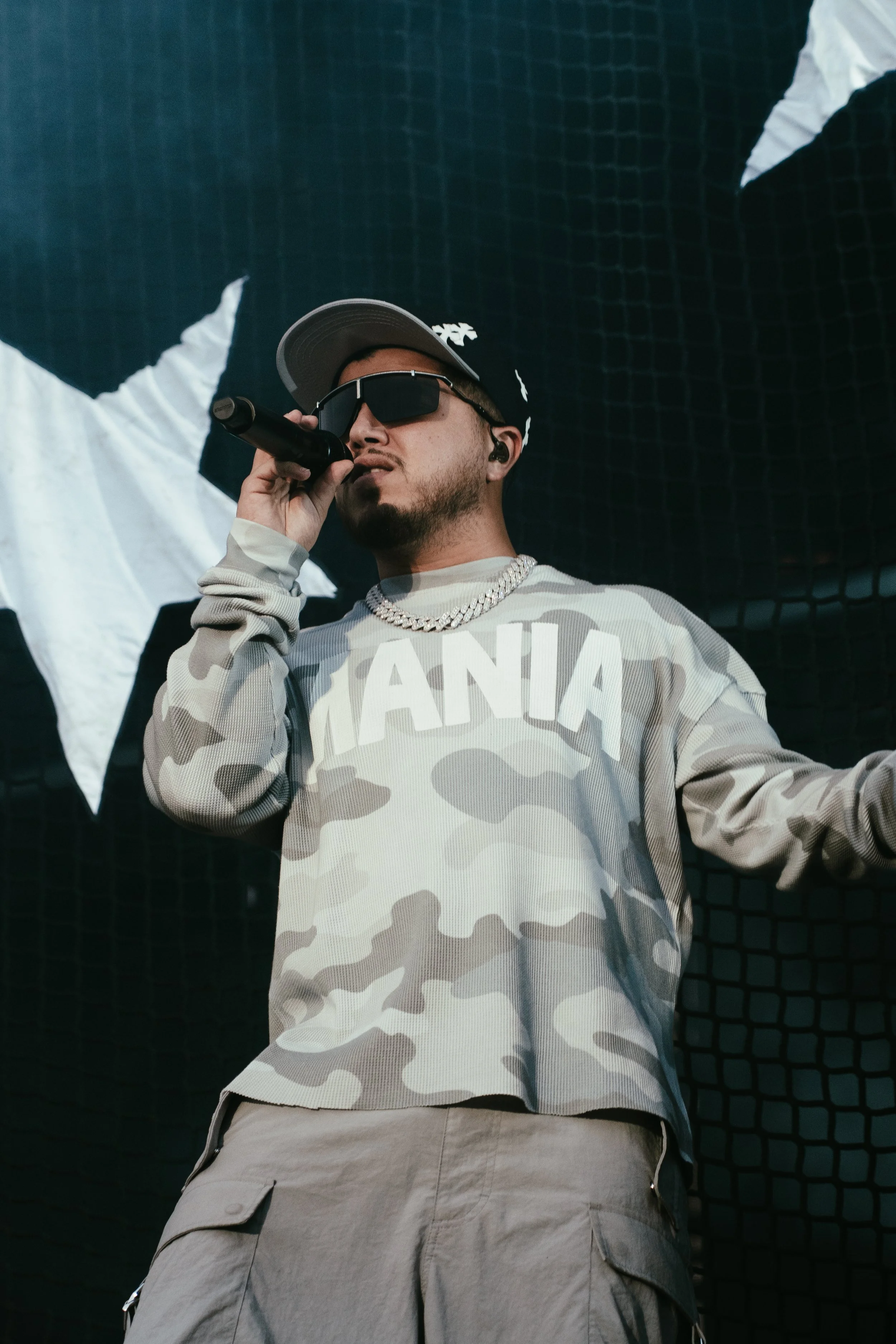 Un rappeur portant des lunettes de soleil, un bonnet, une chaîne en or, un sweat à motifs camouflage avec le mot 'JAMAICA', et un pantalon cargo, en train de performer sur scène, avec une toile de fond noire avec des étoiles blanches.