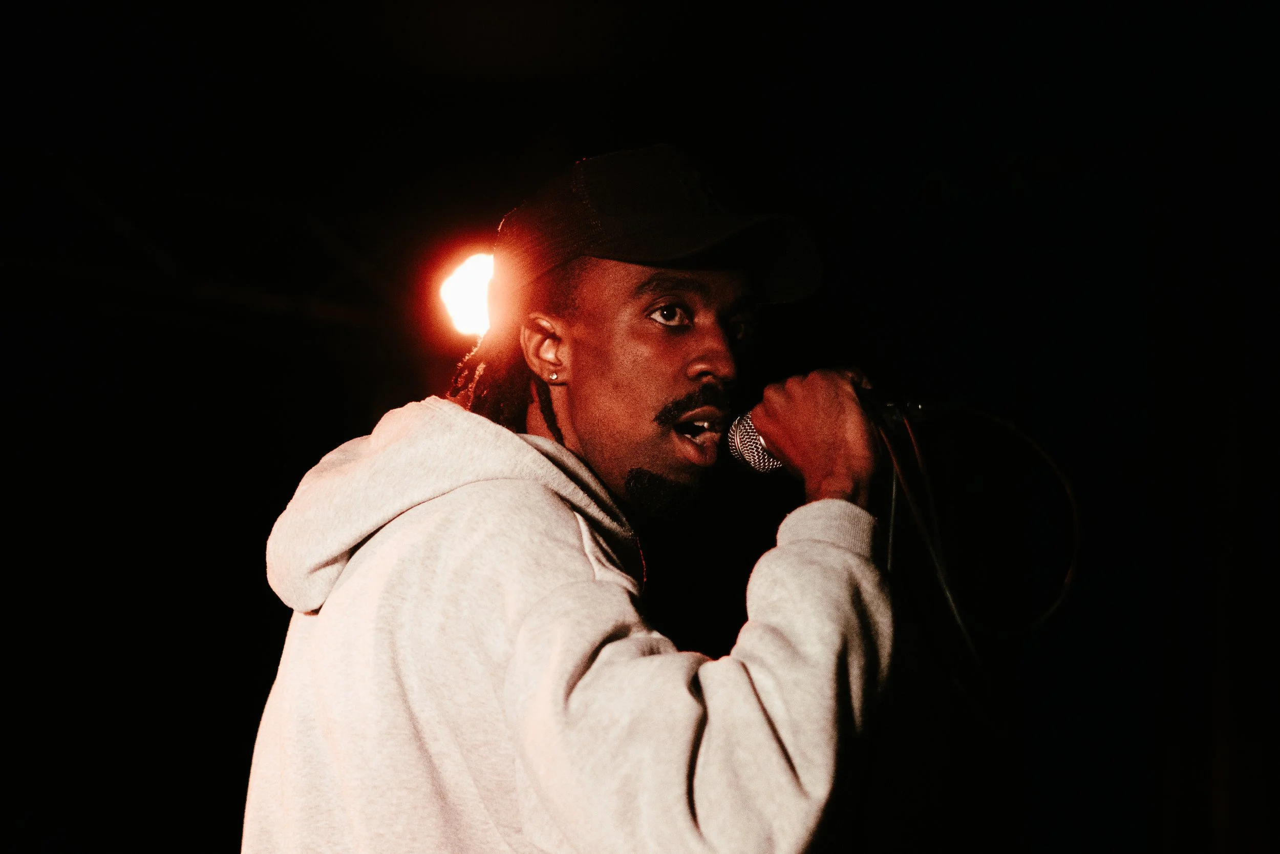 Un homme avec une casquette noire et une veste grise, tenant un microphone, en train de chanter ou de parler dans un environnement sombre, avec une lumière rouge derrière lui.