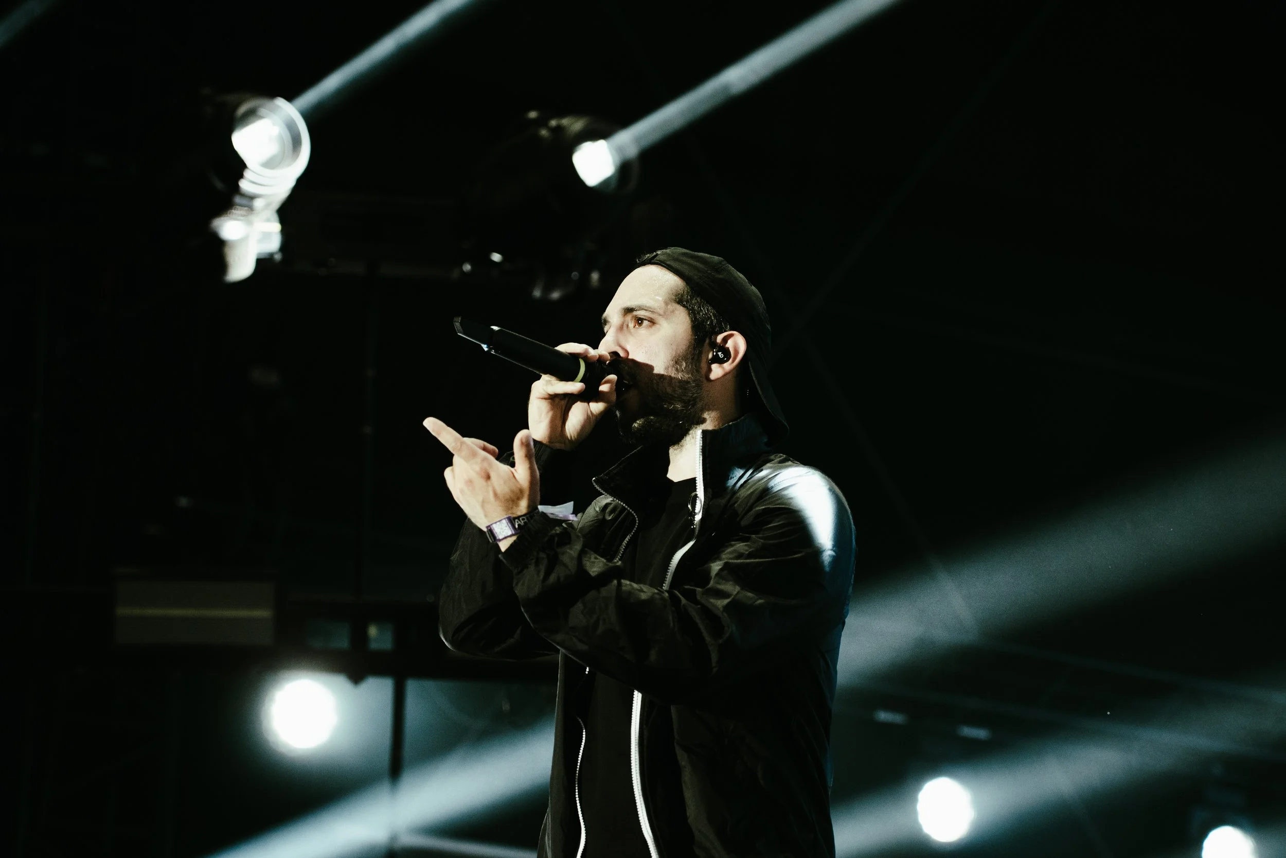Un homme en tenue noire chantant dans un micro sur scène avec des projecteurs en arrière-plan.