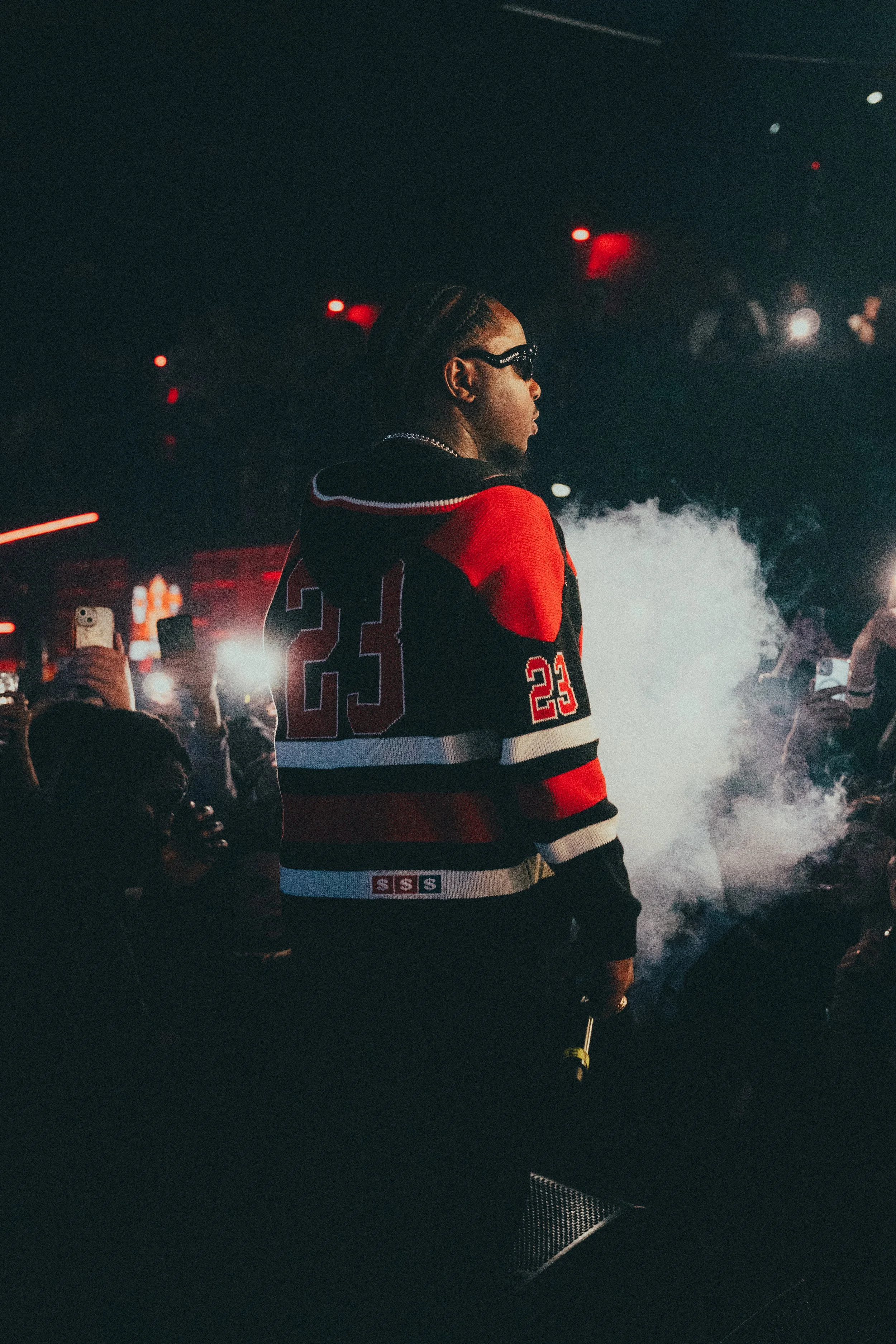 Une personne portant un maillot de hockey rouge, noir et blanc avec le numéro 23, se tient dans une ambiance sombre avec de la fumée, entourée de personnes prenant des photos.