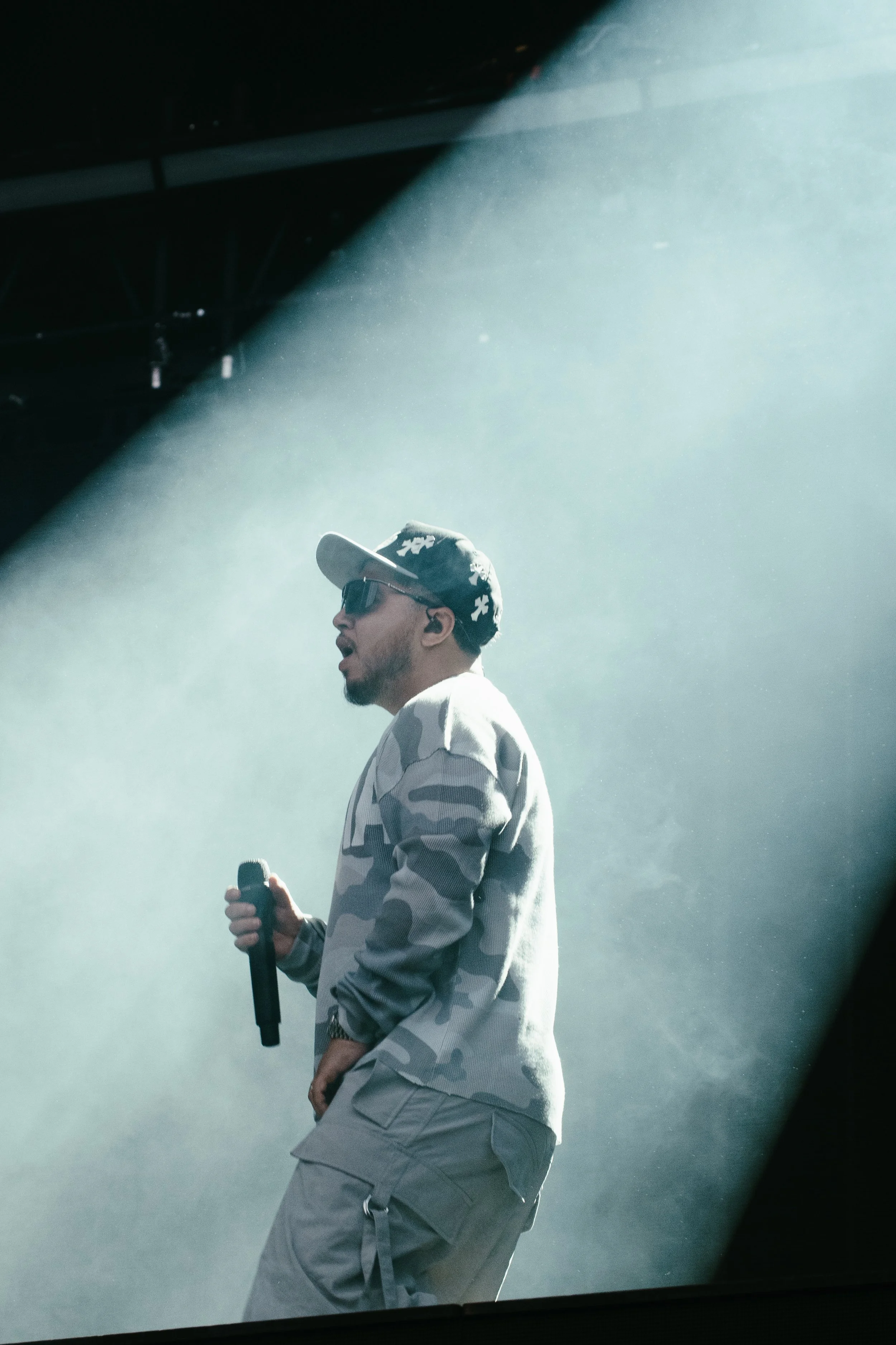 Un homme portant un chapeau, des lunettes de soleil, un sweatshirt camouflage, et des pantalons cargo, tenant un microphone en scène, éclairé par un faisceau lumineux.