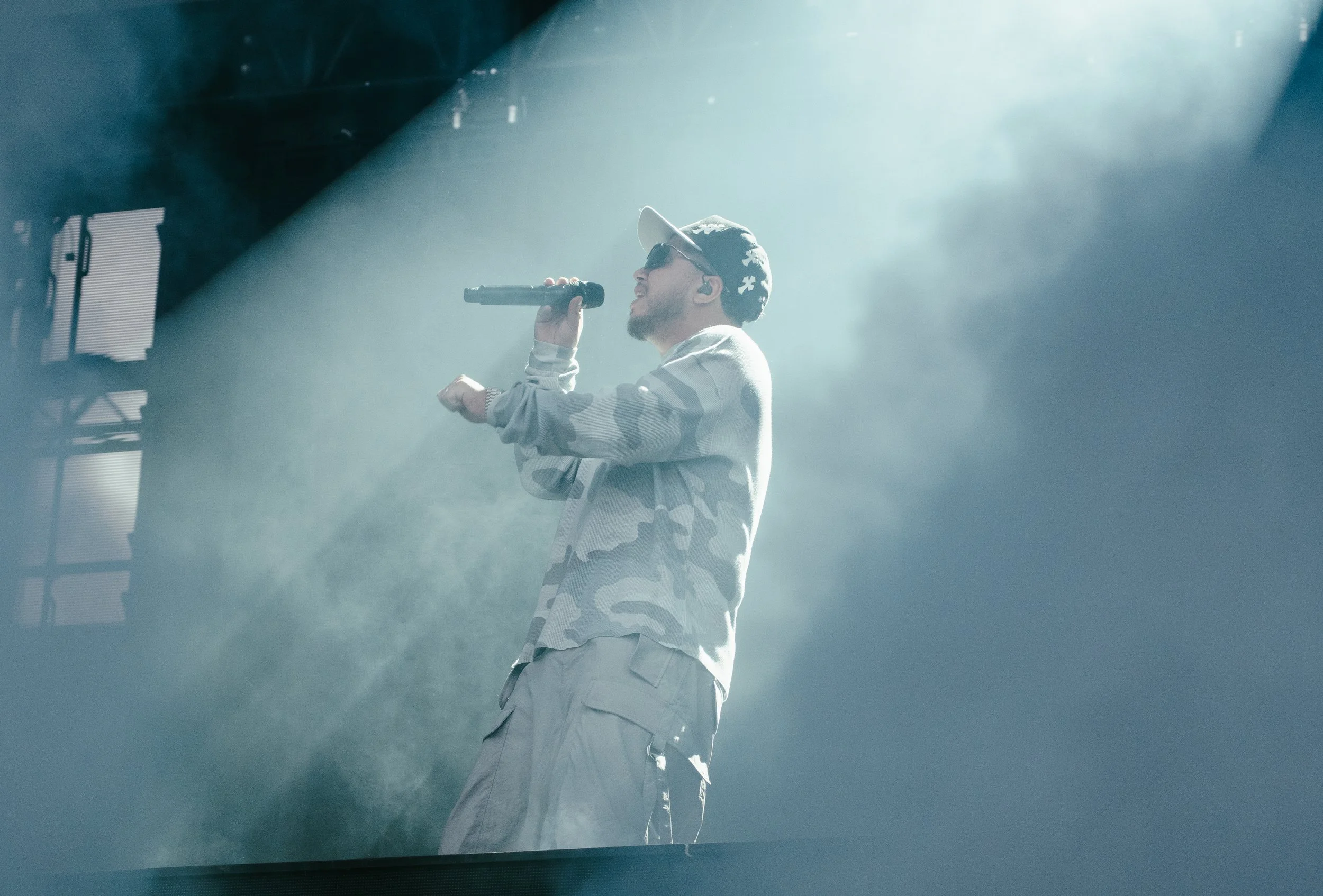 Un homme portant une chemise à motifs camouflage, un pantalon cargo, une casquette et des lunettes de soleil, chante dans un microphone sur scène avec des effets lumineux et de la fumée.
