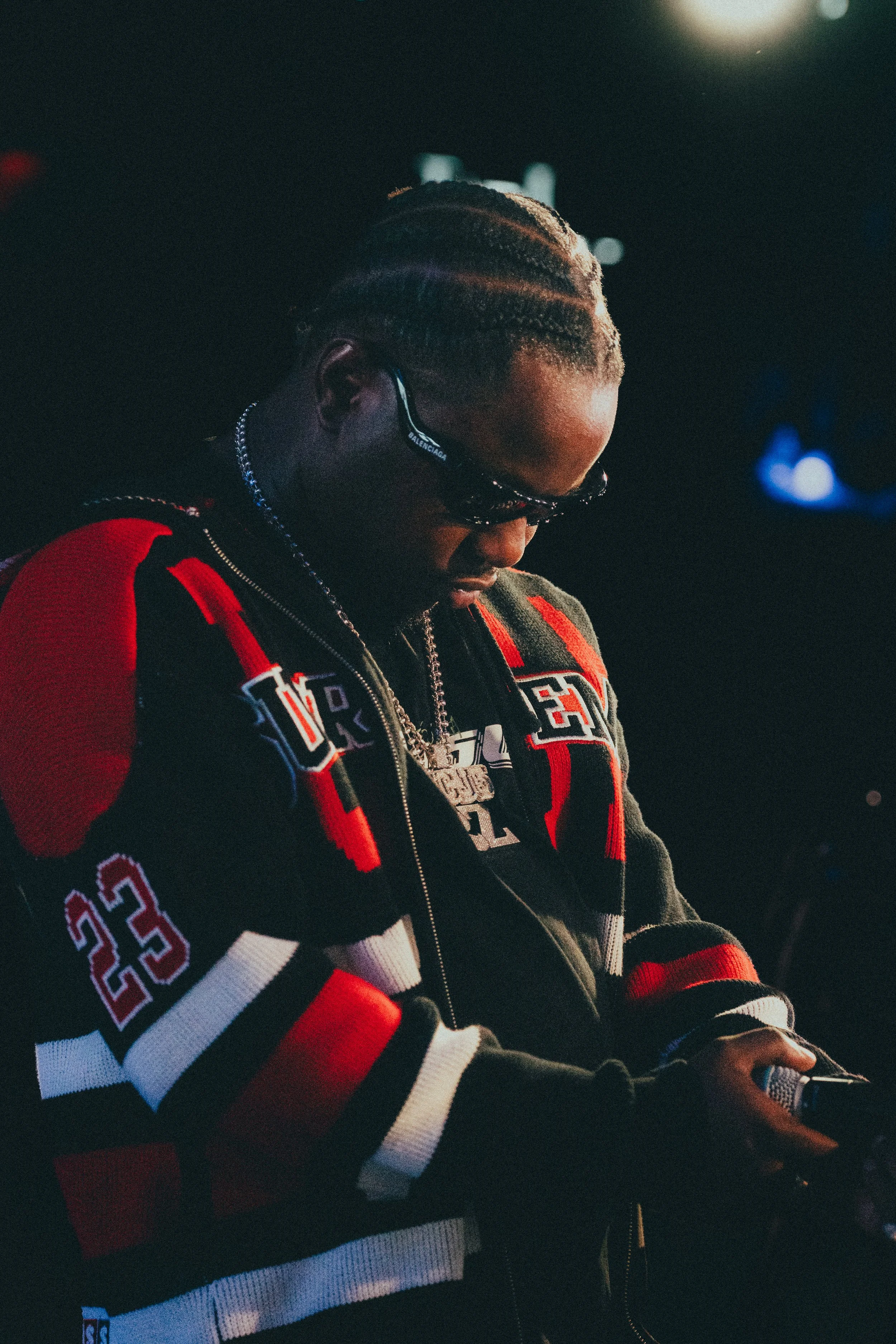 Homme avec des dreadlocks portant des lunettes de soleil, un blouson noir et rouge avec le numéro 22, et un collier en chaîne, regardant son téléphone dans un environnement sombre et éclairé par des lumières