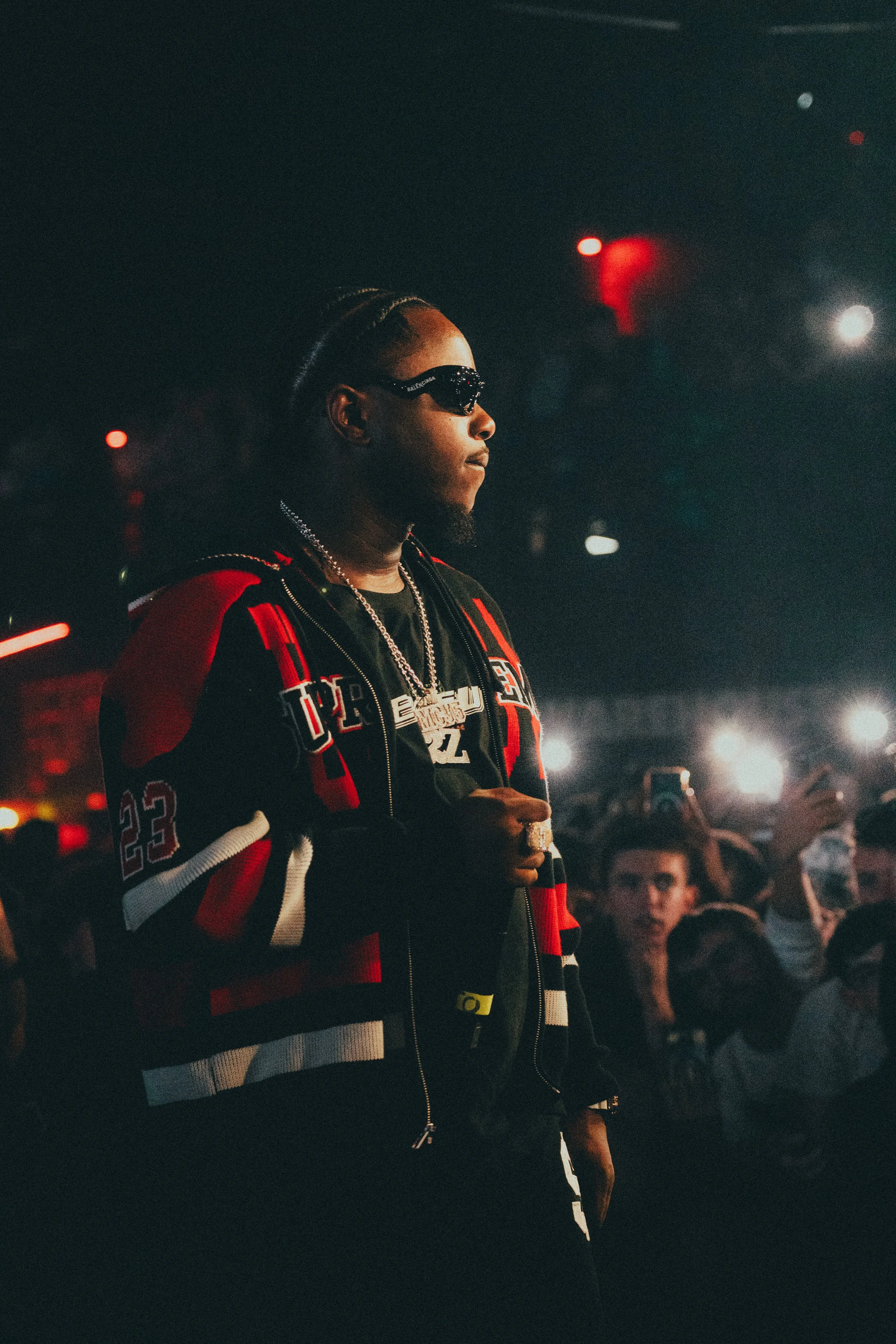 Un homme portant des lunettes de soleil, un sweat à capuche noir et une veste noire et rouge, avec des bijoux, dans une foule lors d'un concert ou événement en intérieur avec des lumières et des écouteurs.