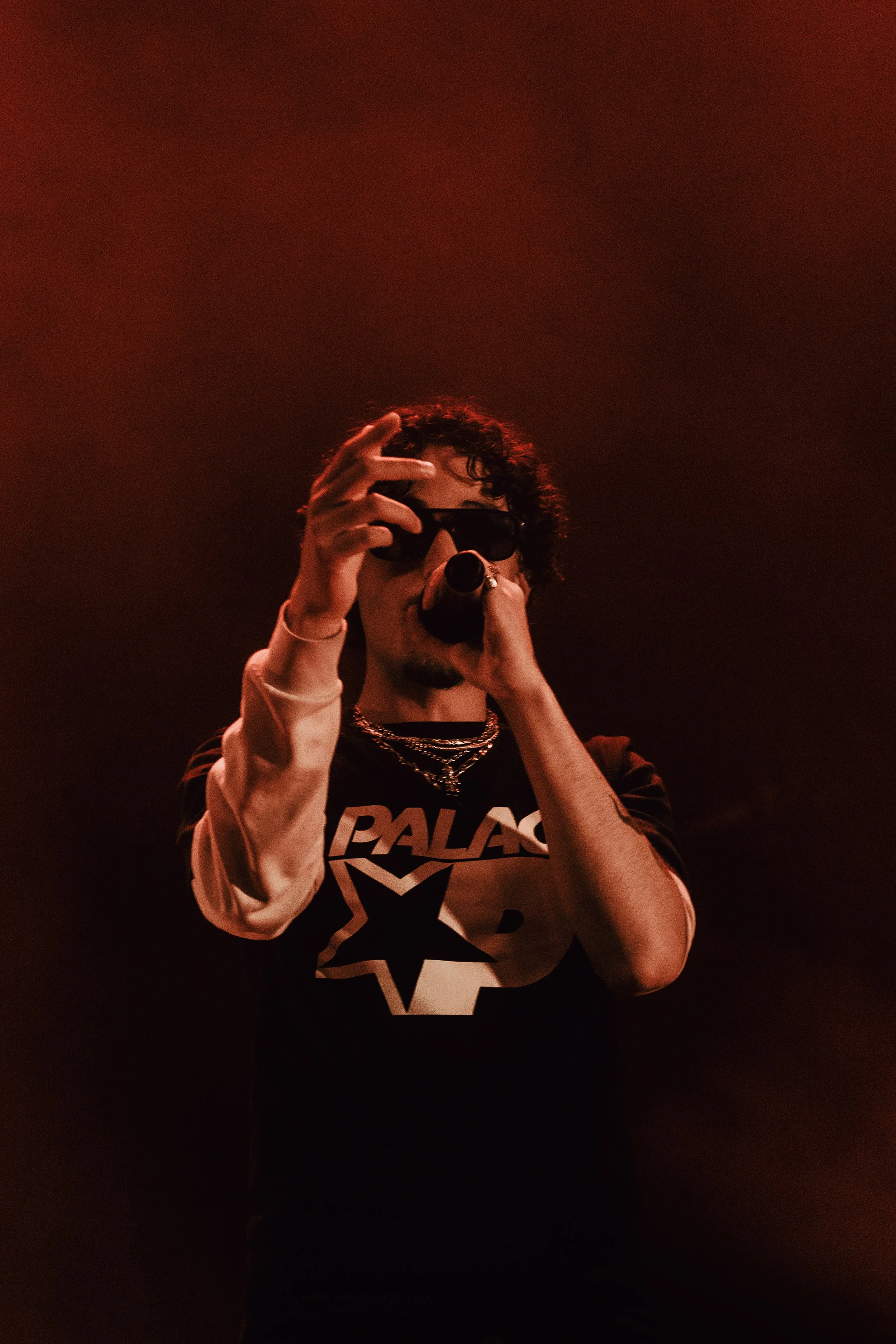 Une personne portant des lunettes de soleil, un t-shirt avec un design en étoile et des chaînes, tenant un micro, photographiée dans un éclairage rouge.