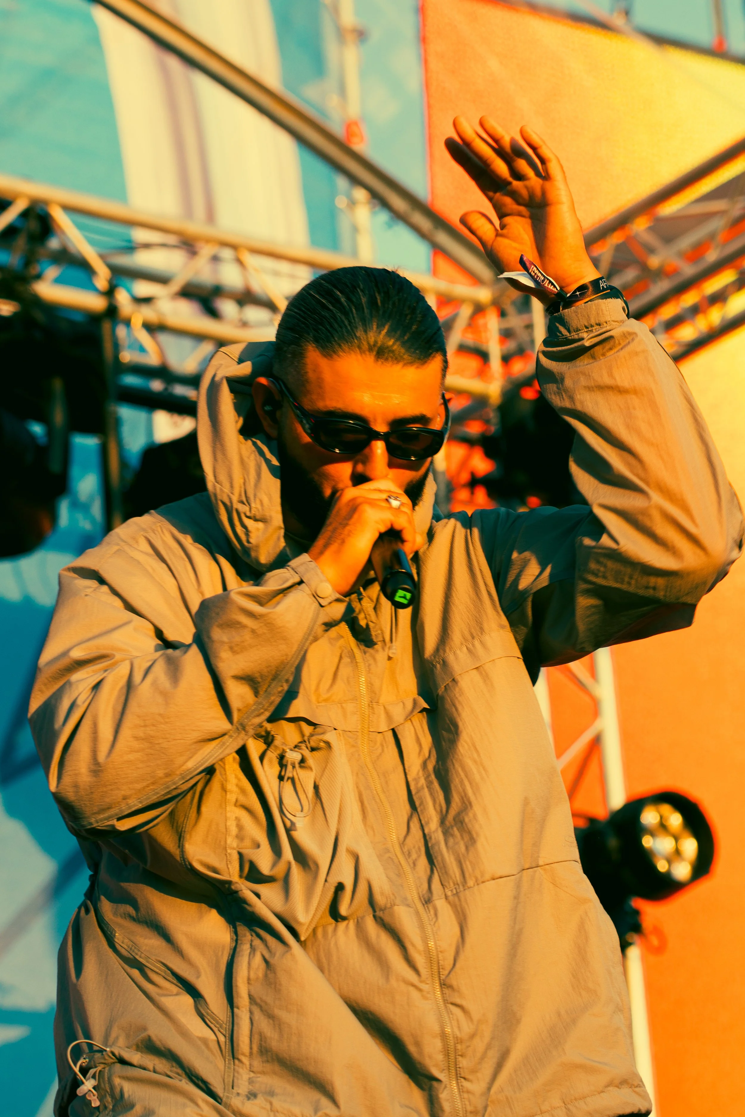 Un homme portant des lunettes de soleil, un blouson beige, tenant un microphone, en train de chanter ou de parler sur scène lors d'un événement en plein air.