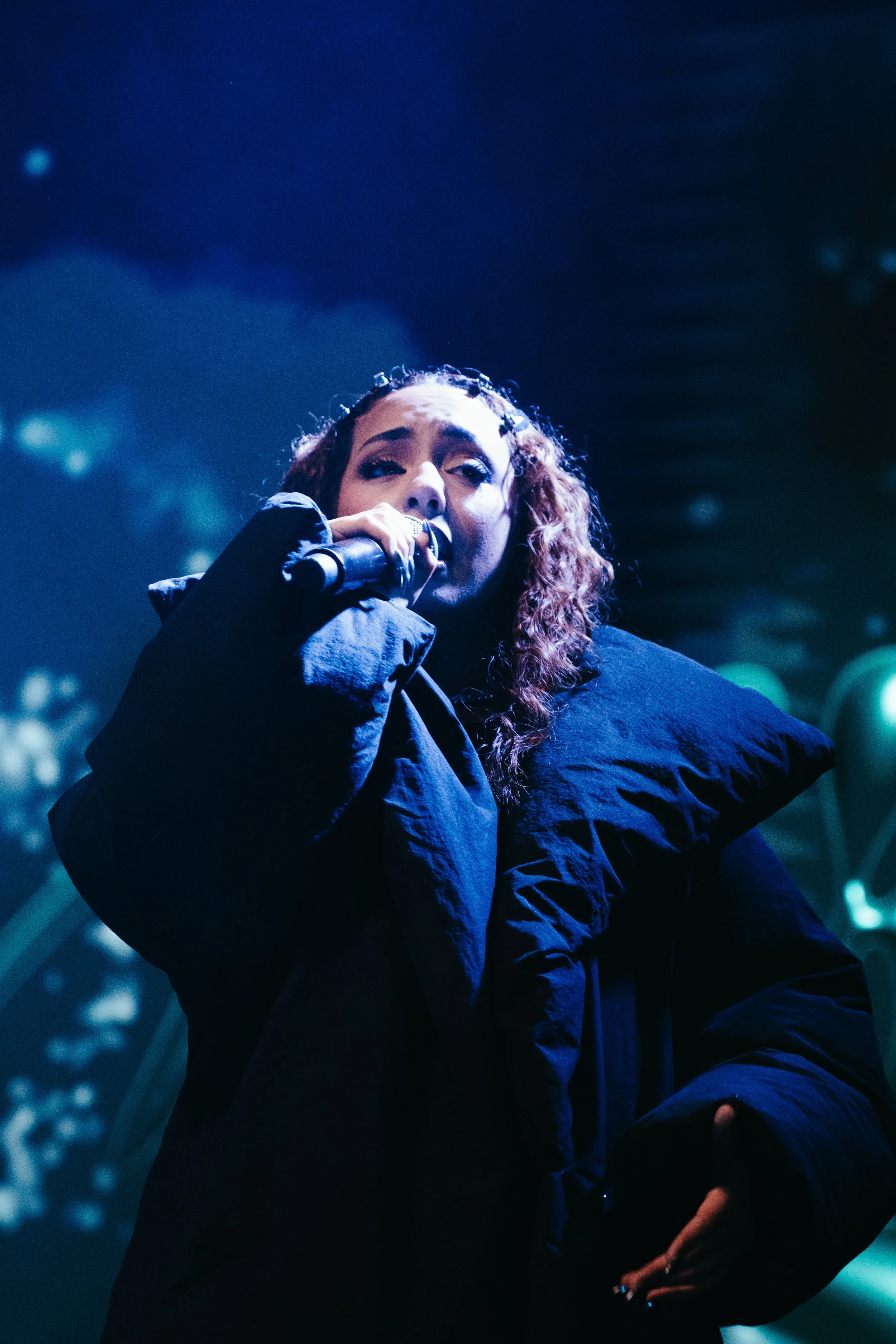Une femme chante dans un cadre sombre avec un micro, portant un vêtement noir avec des épaules bouffantes, éclairée par des lumières bleues et vertes.