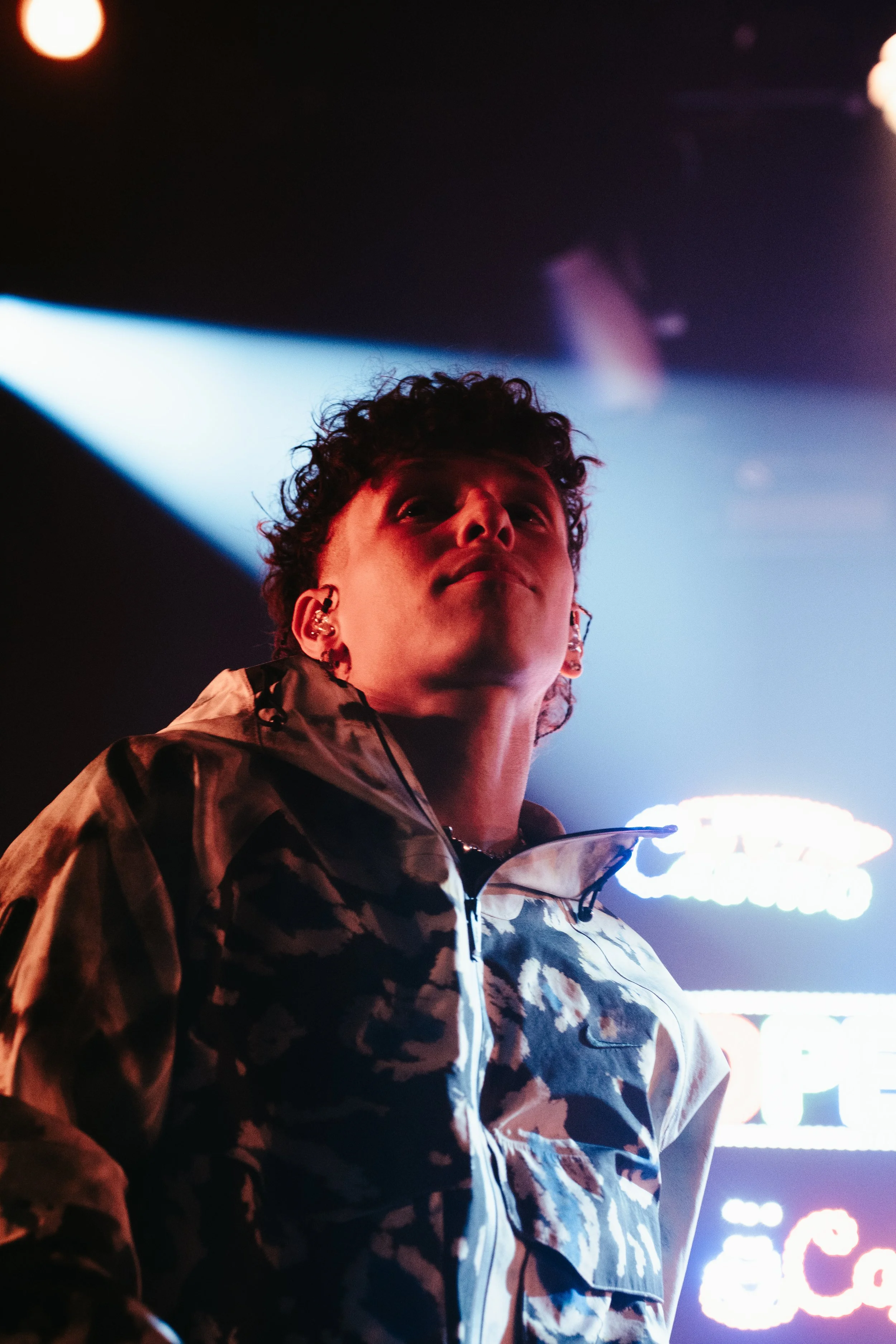 Jeune homme dans la lumière artificielle, portant un blouson camouflage, dans un environnement sombre avec des néons