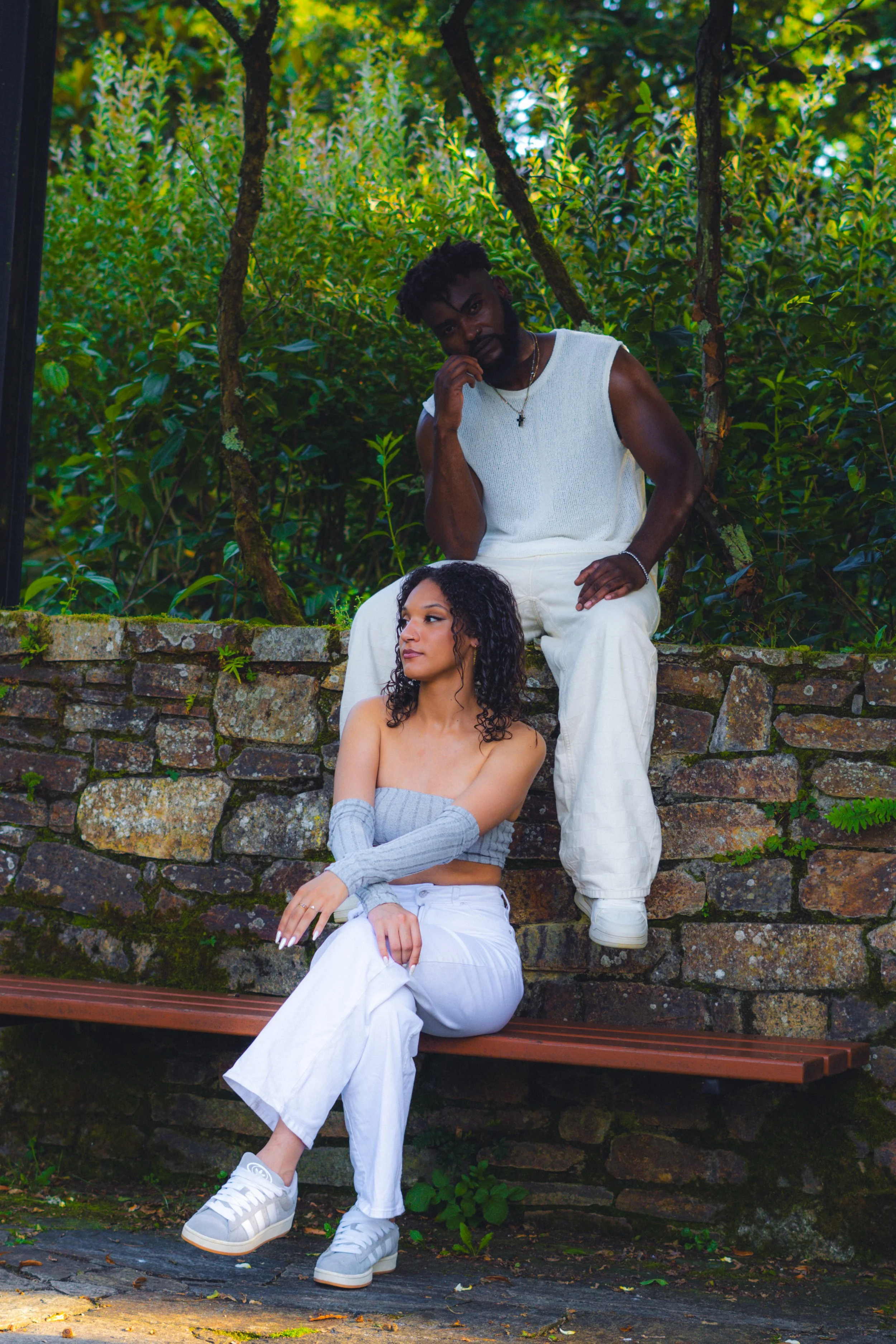 un homme et une femme posent côte à côte devant un mur en pierre dans un espace vert. L'homme est assis sur le mur, portant un vêtement blanc sans manches et des pantalons blancs, avec une touche de style en pendentif. La femme est assise sur un banc