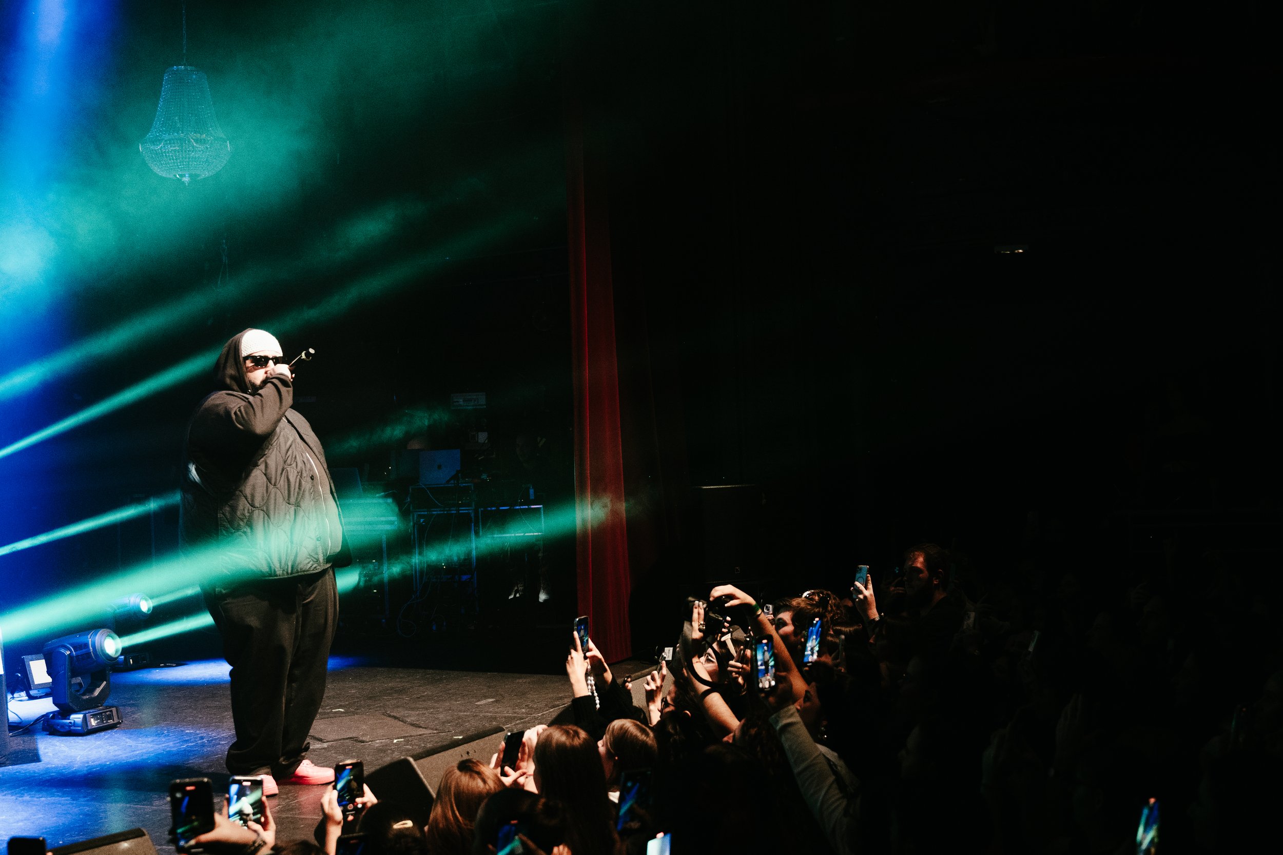 Un artiste portant des lunettes de soleil et une capuche chante sur scène avec des lumières vertes et bleues, face à une foule de spectateurs qui prennent des photos avec leurs téléphones.