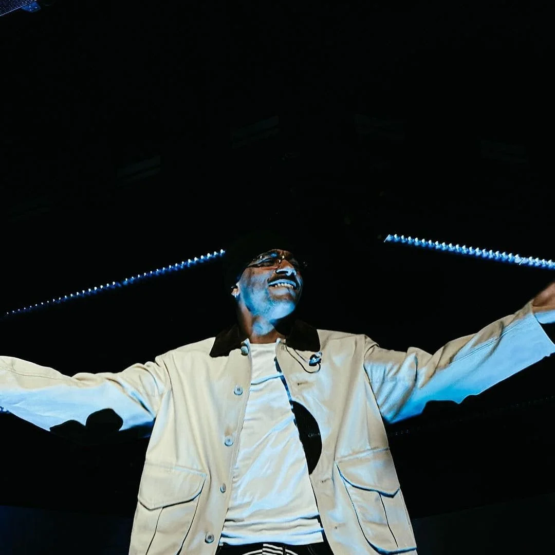 MISTER V souriant, portant des lunettes et une veste beige, avec ses bras ouverts, dans un environnement sombre avec des lumières bleues.