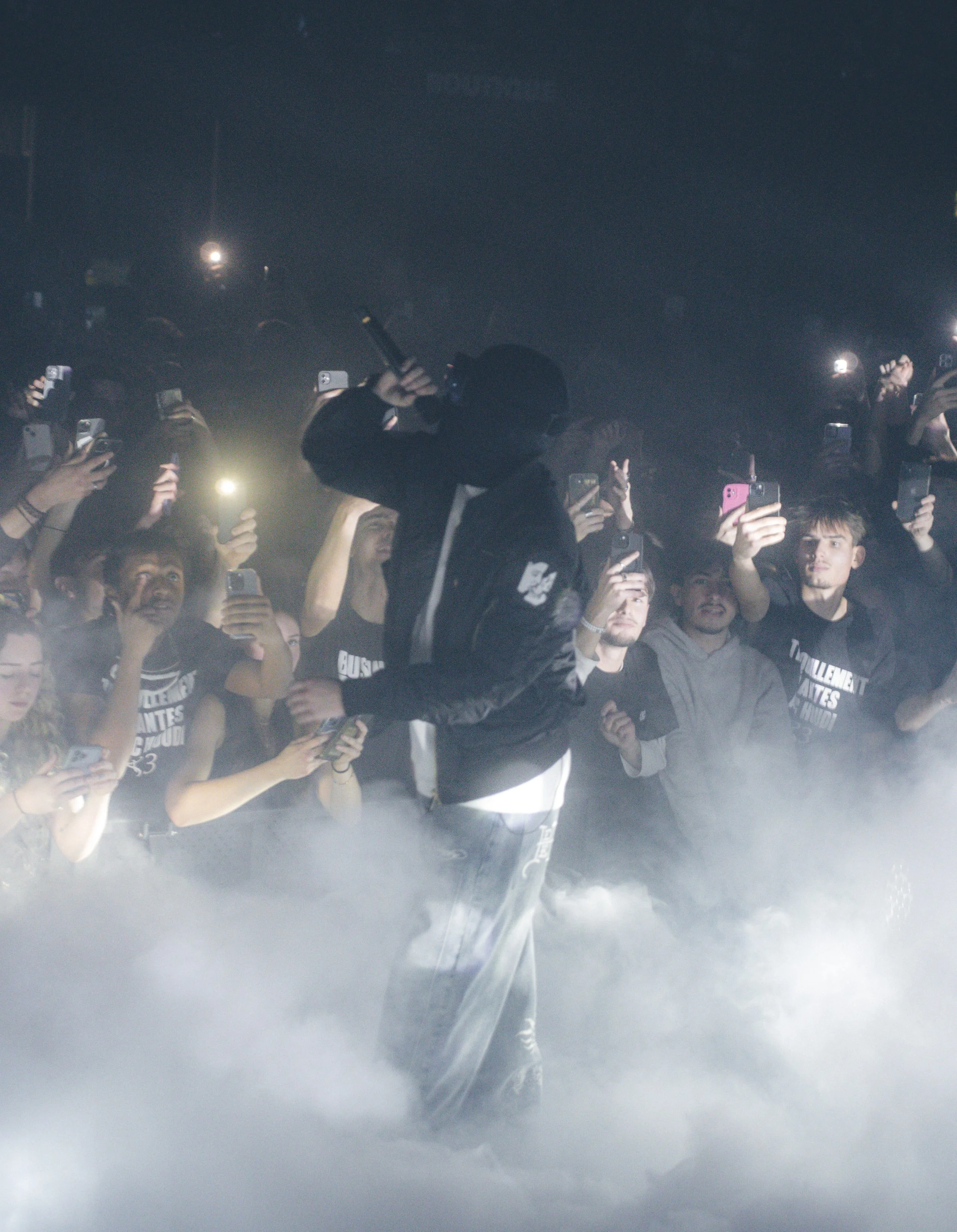 Une personne portant une veste noire et un casque, tenant un microphone, se dresse au centre entourée de personnes qui prennent des photos avec leurs téléphones dans une ambiance sombre et brumeuse.