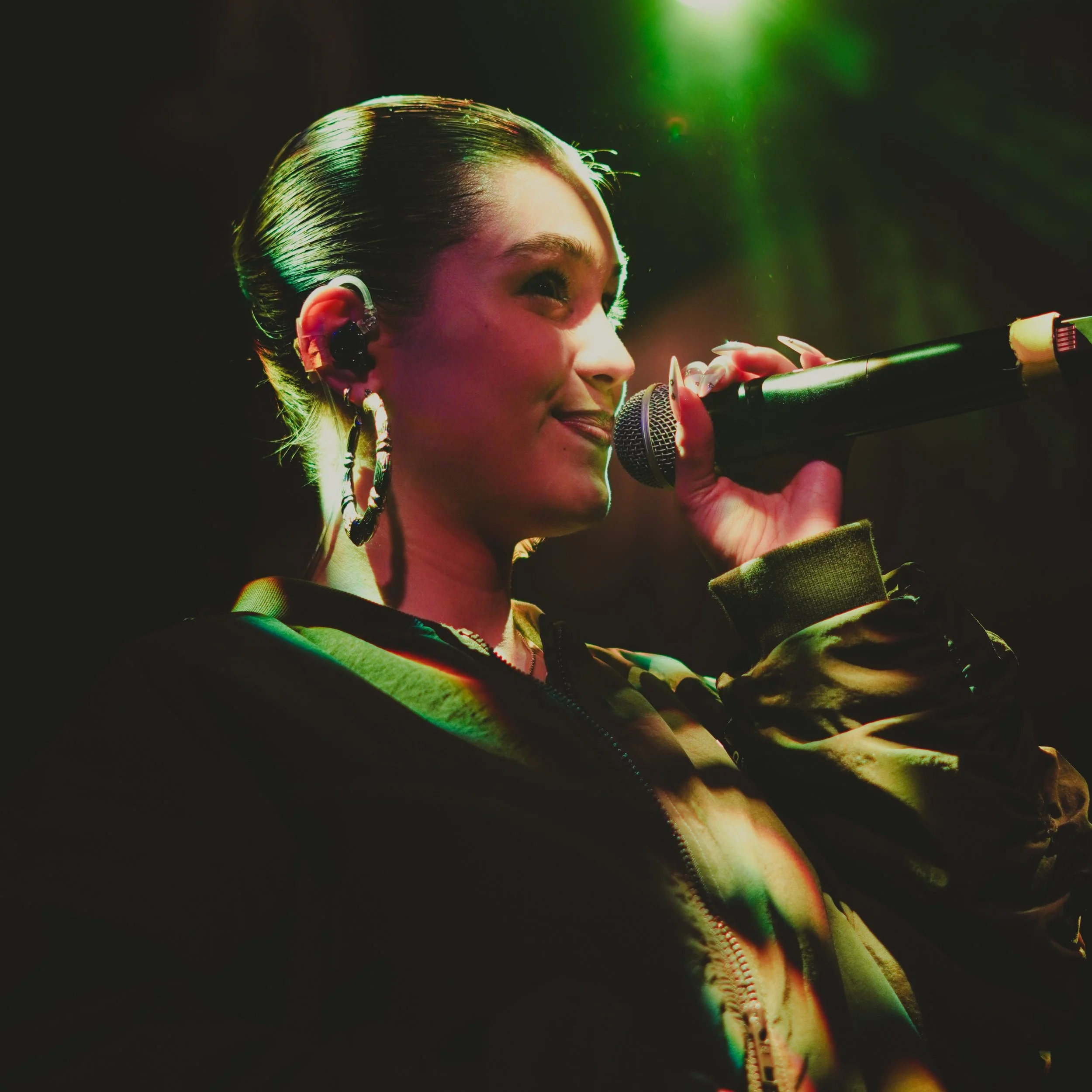 Une femme qui chante ou parle dans un micro, avec des lumières colorées vertes et roses, portant des bijoux et une veste textile.